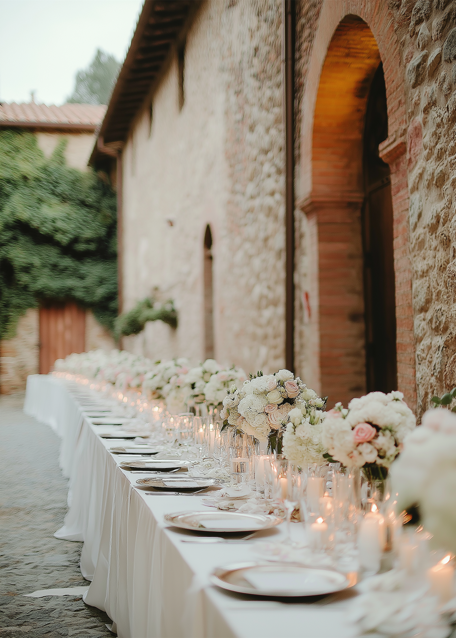 Beachside wedding reception setup with a wooden table decorated with greenery and white linens, and an aisle leading to a floral arch over the sand with ocean views.