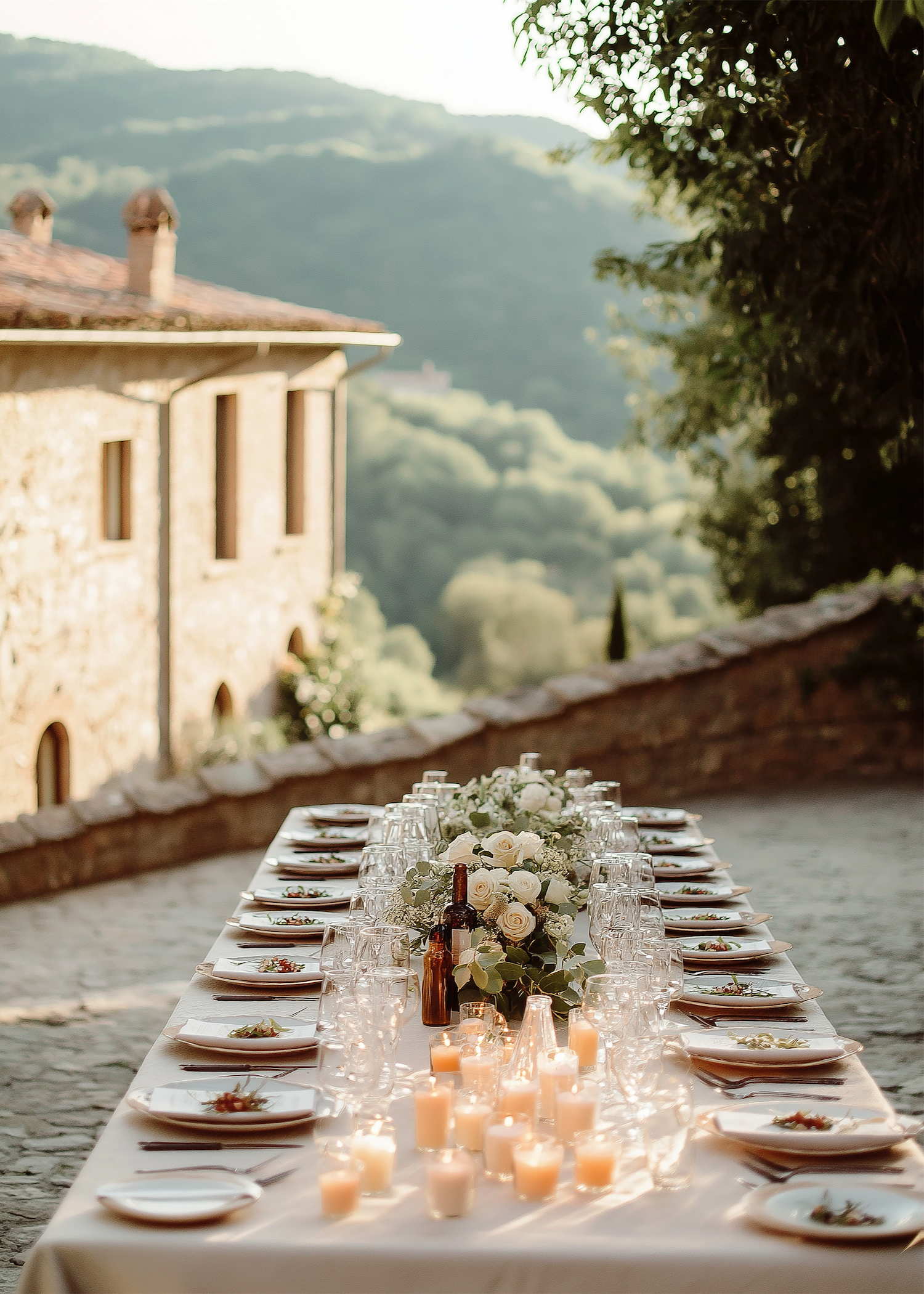 Long banquet table decorated with white and pink flower arrangements, candles, and glassware, set outdoors against a rustic stone and brick building.