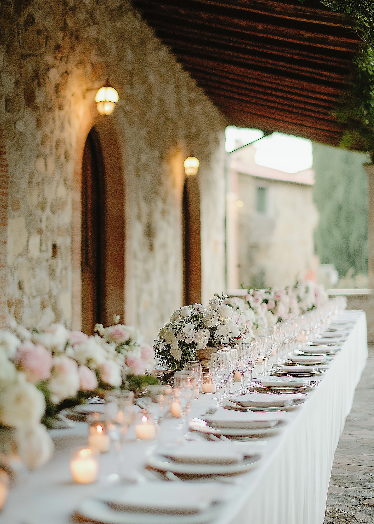 Long banquet table decorated with white floral arrangements and candles, set for a formal event outdoors against a stone wall with arched windows and warm lighting.