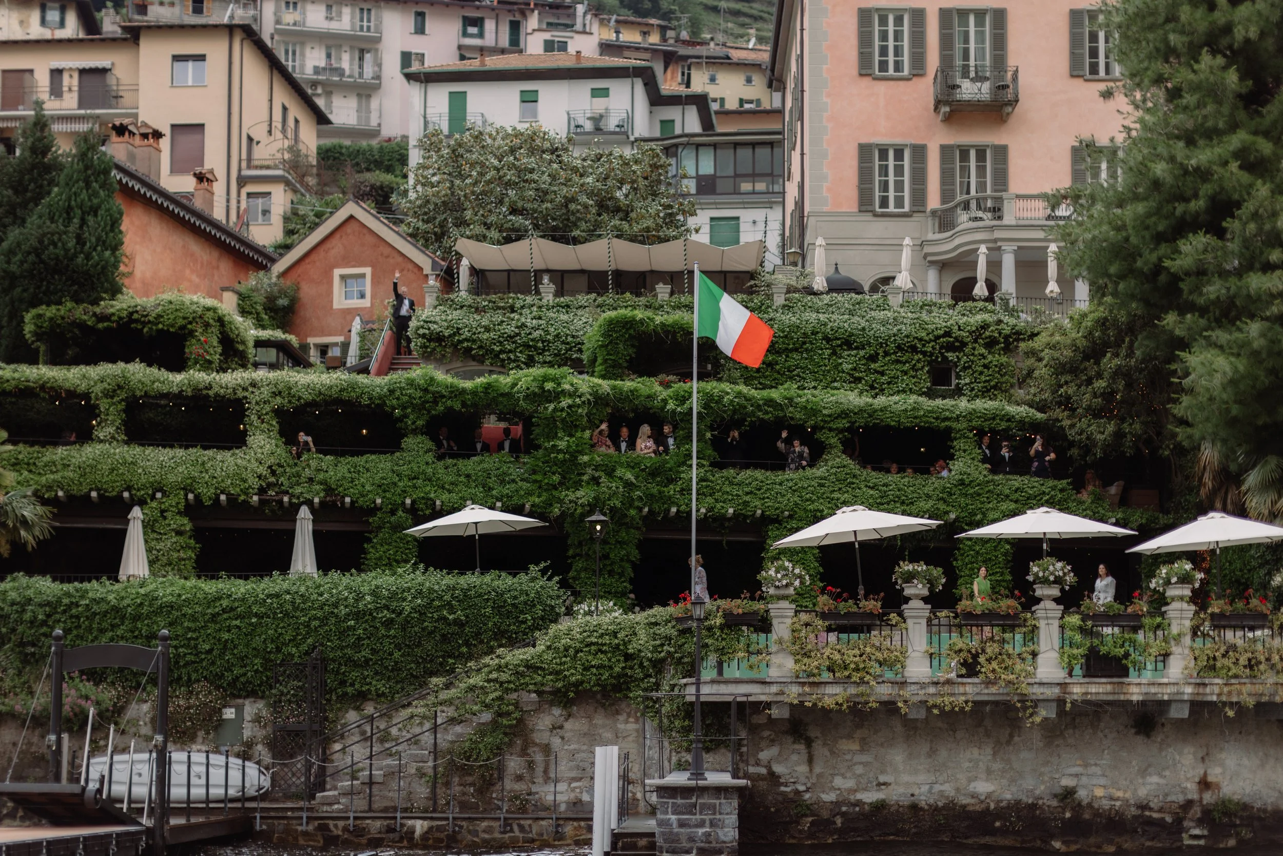 A multi-level outdoor terrace with greenery, umbrellas, and people dining, featuring an Italian flag in the center, hillside houses in the background, and a waterfront at the bottom.