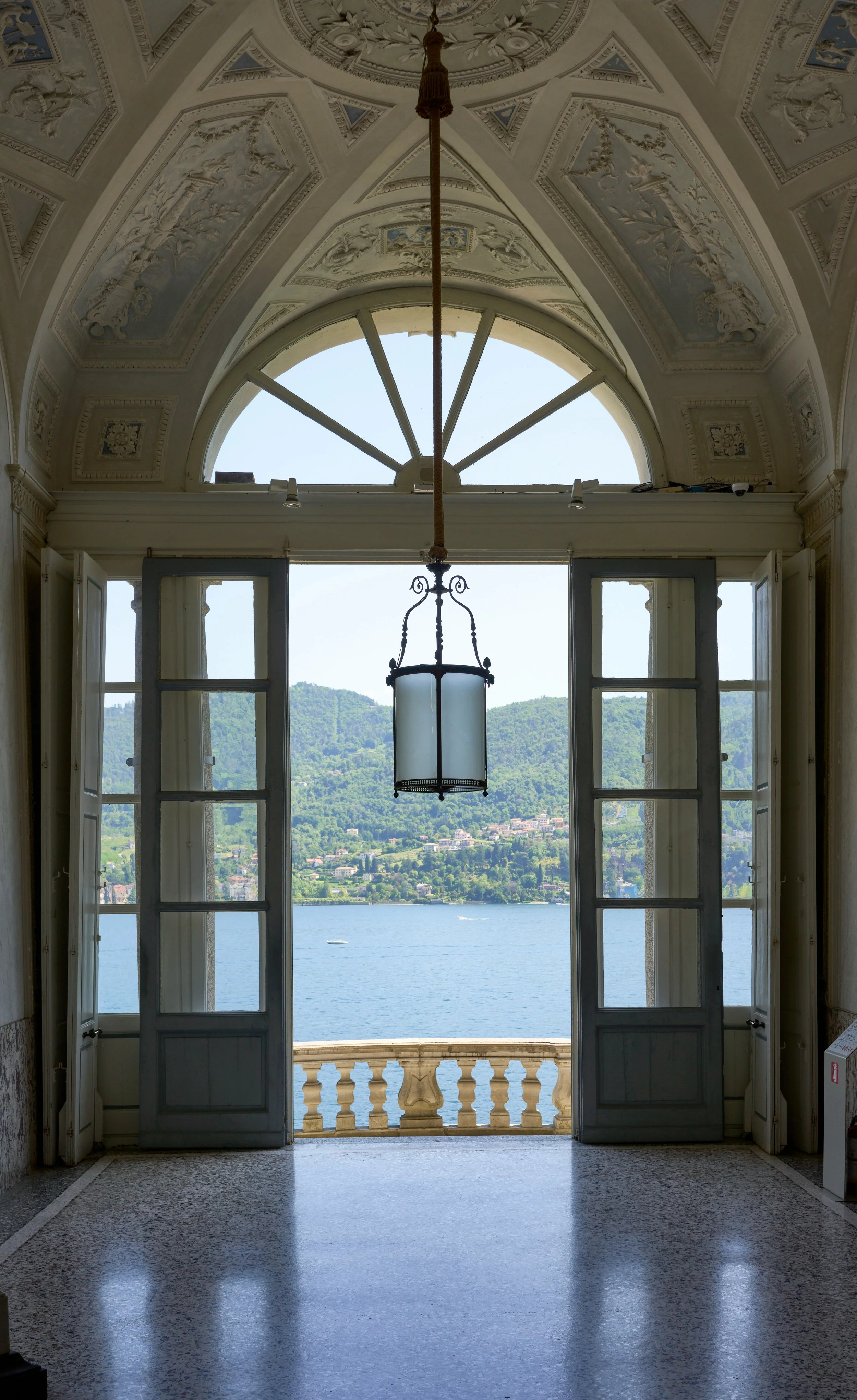 Open double doors of an elegant building overlooking a lake and green hills with scattered houses and trees.