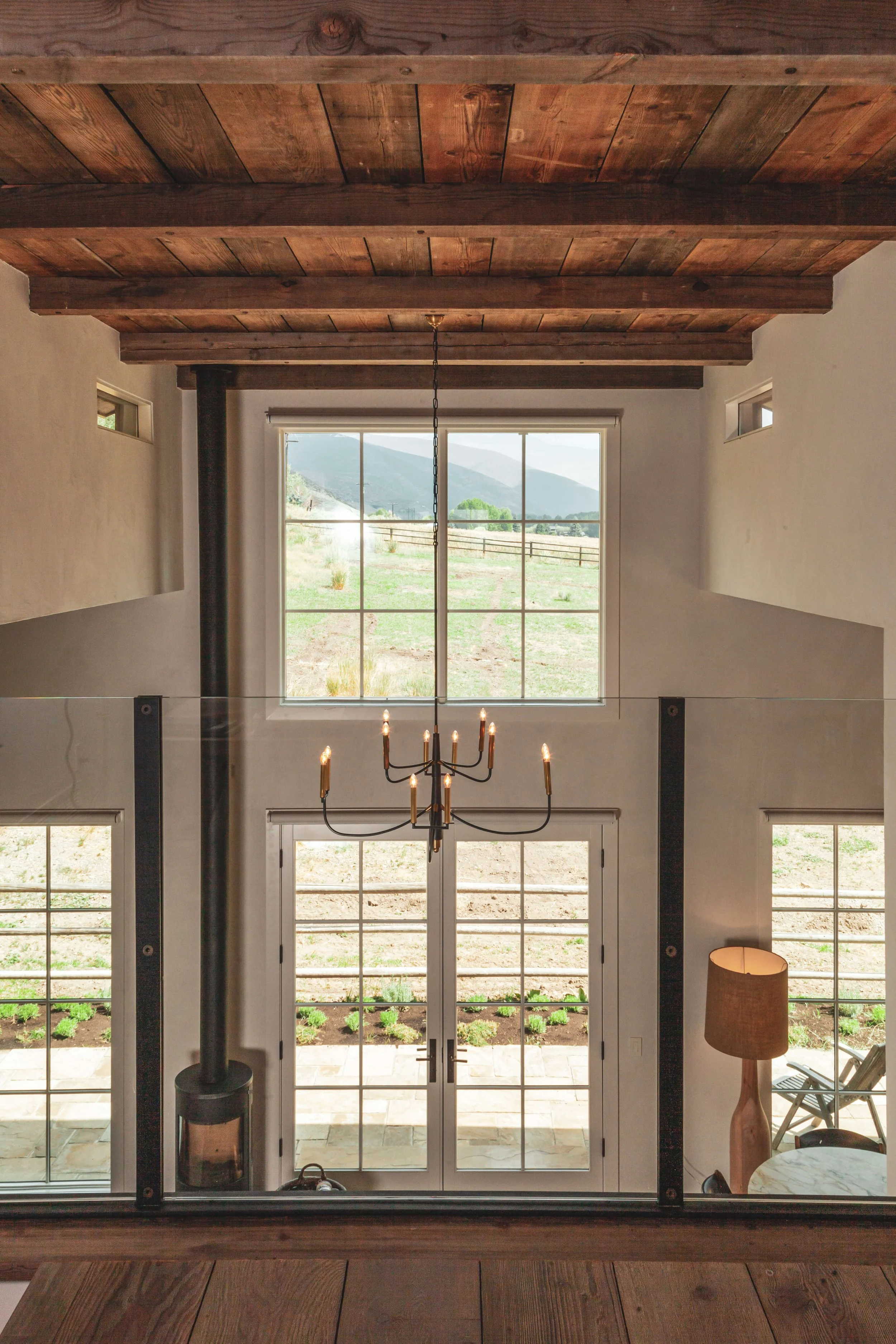 Interior view of a room with large glass doors and windows showing a scenic mountain landscape outside. There is a black wood-burning stove on the left, a modern chandelier hanging from a wooden ceiling, and a table with a lamp and a chair on the rig
