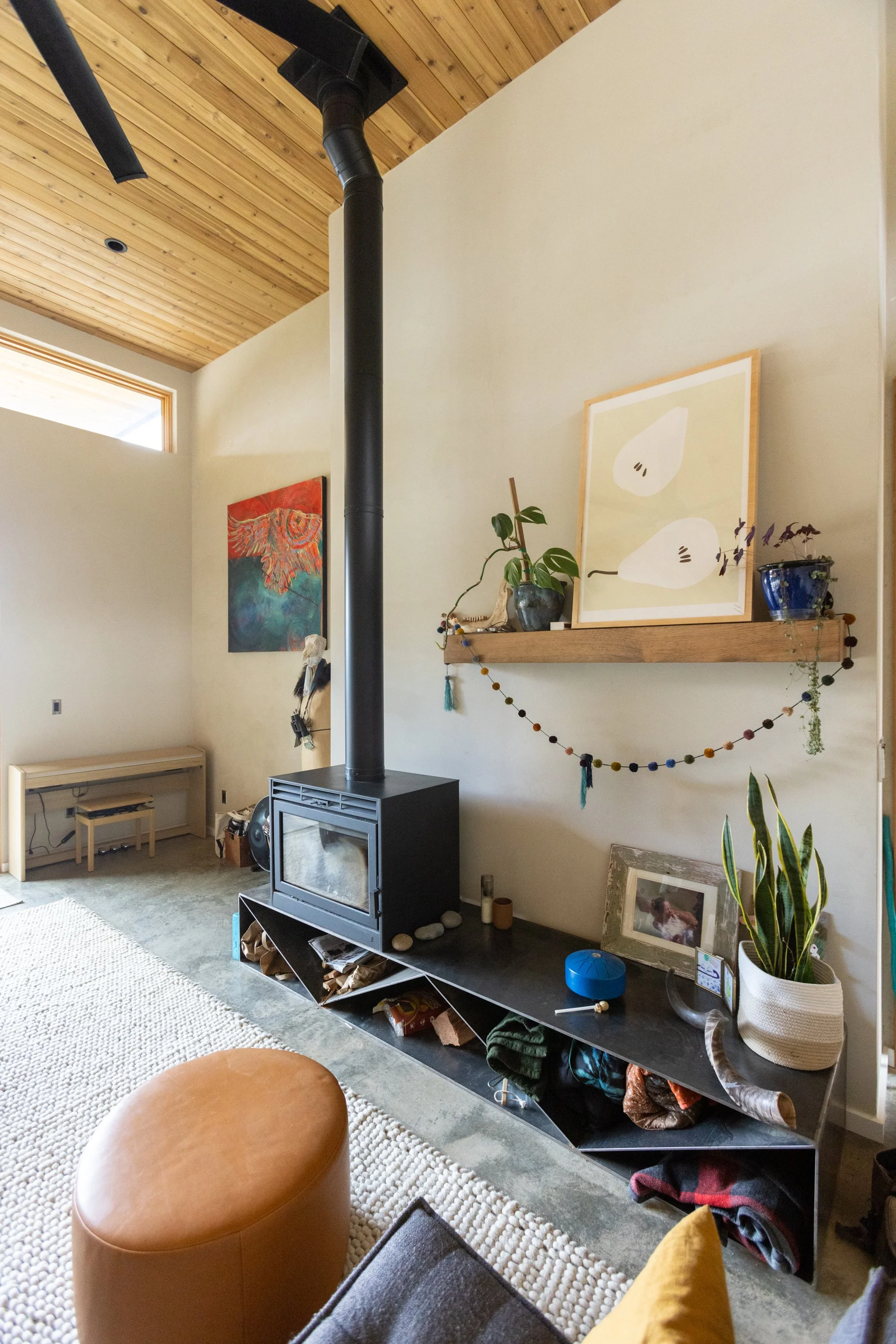 A living room with a wood stove, potted plants, framed artwork, a beige carpet, and a leather ottoman, with a wooden ceiling and light-colored walls.