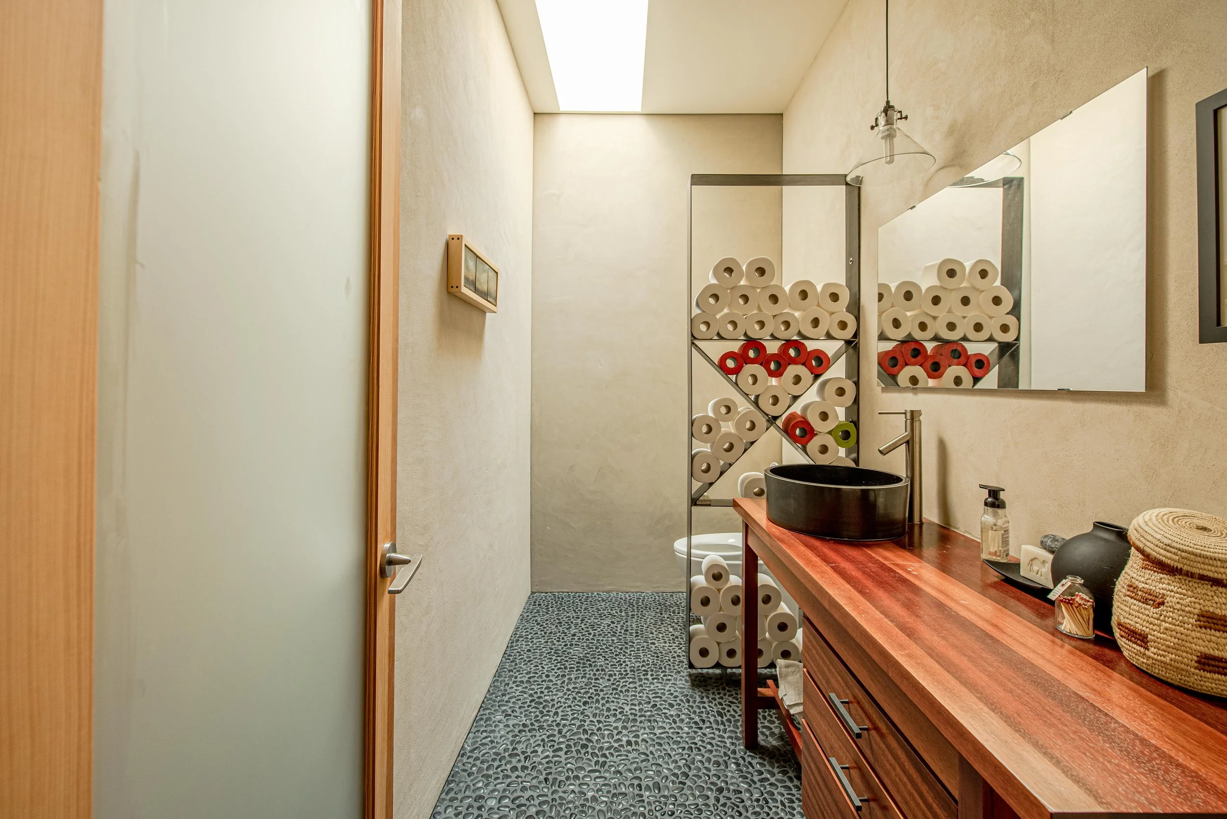 A bathroom with a wooden vanity, black vessel sink, mirror, and a metal towel rack filled with toilet paper rolls, with a small shower area in the background.