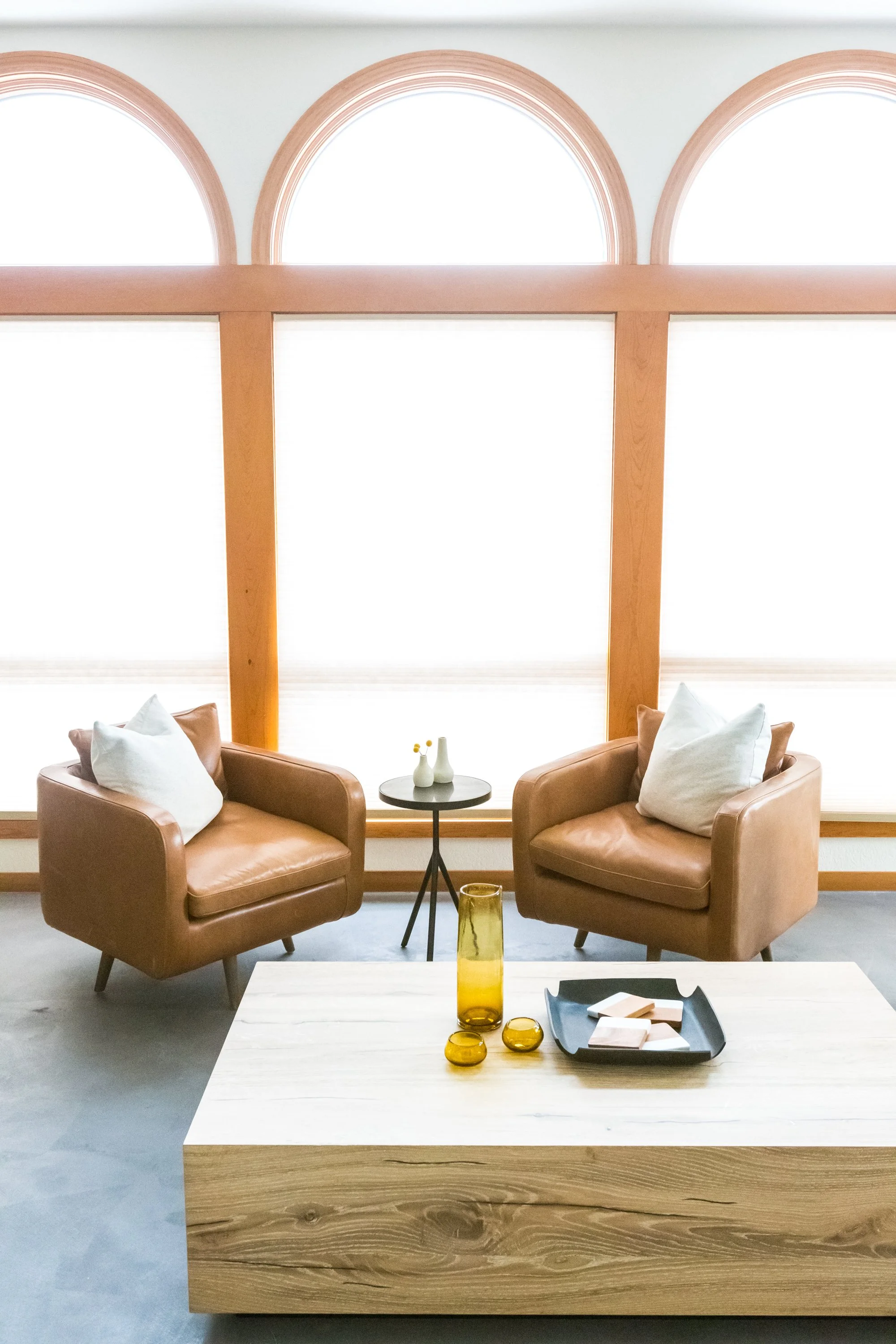 Living room scene with two tan leather armchairs, a small black side table with vases, and a large white coffee table with a black tray, yellow decorative objects, and a wood panel at the front, in front of three large arched windows with wooden trim