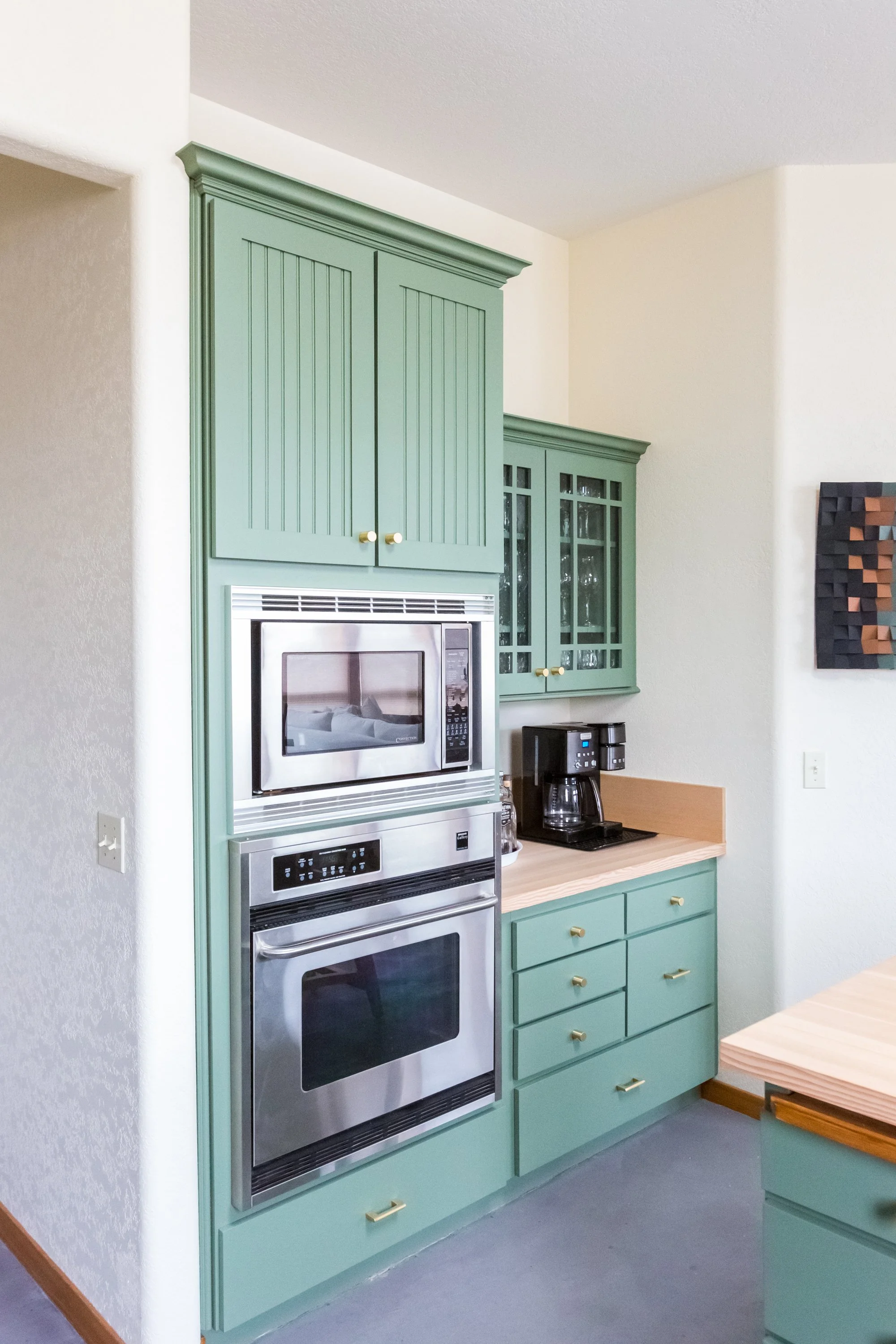 Kitchen with mint green cabinets, a stainless steel microwave and oven, black coffee maker, and a wooden table with green cabinets underneath.