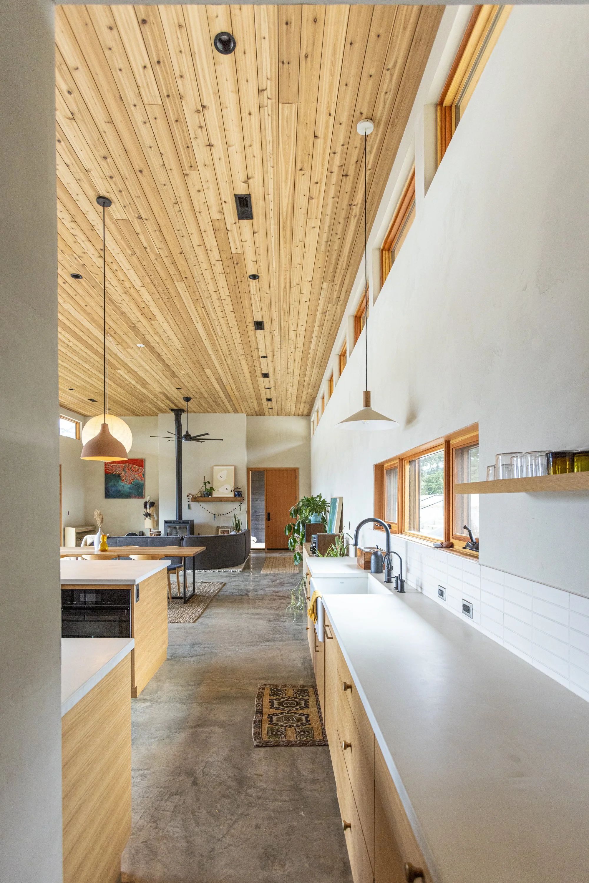 Open-concept kitchen and living space with a wooden ceiling, white walls, and a large window. The kitchen features a white countertop, wooden cabinets, a sink, and shelves with glasses. The living area includes a sofa, a colorful painting, and a wood