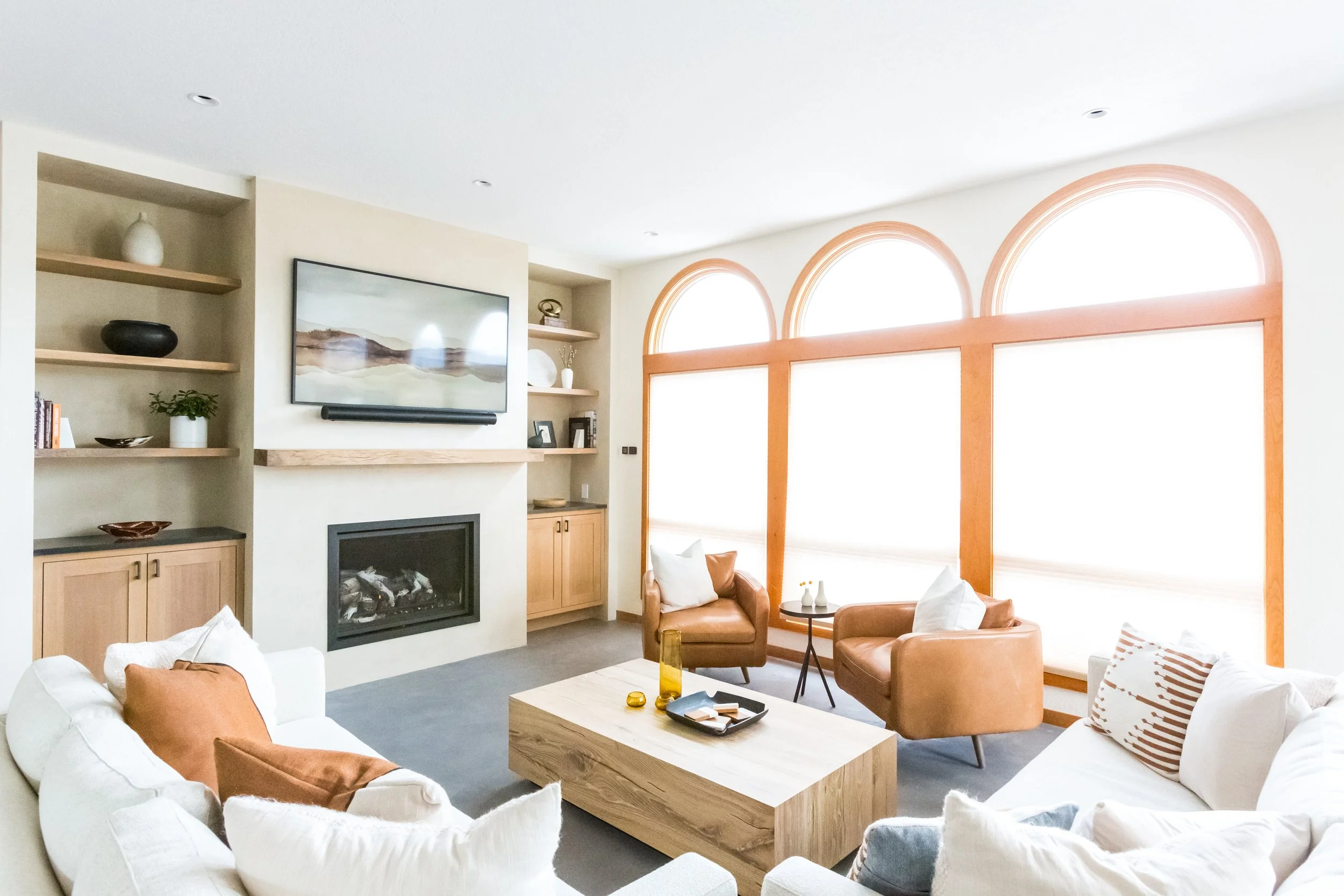 Living room with white sofas, brown leather chairs, a wooden coffee table, a large window with arched frames, a wall-mounted TV above a fireplace, open shelves with decor, and decorative items on tables.