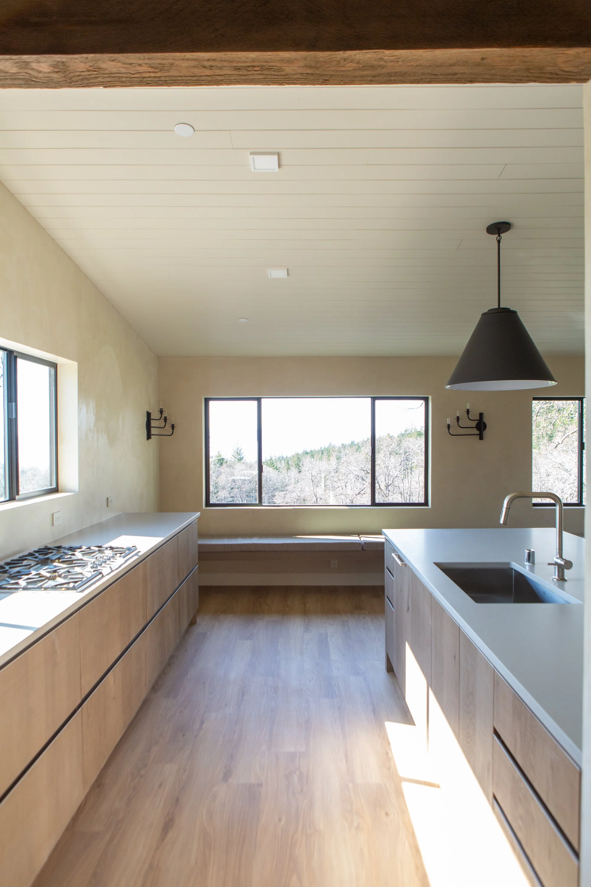 Modern kitchen with light wood cabinets, a white countertop with an undermount sink, black faucet, stove, large windows, a black hanging pendant light, and wood flooring.