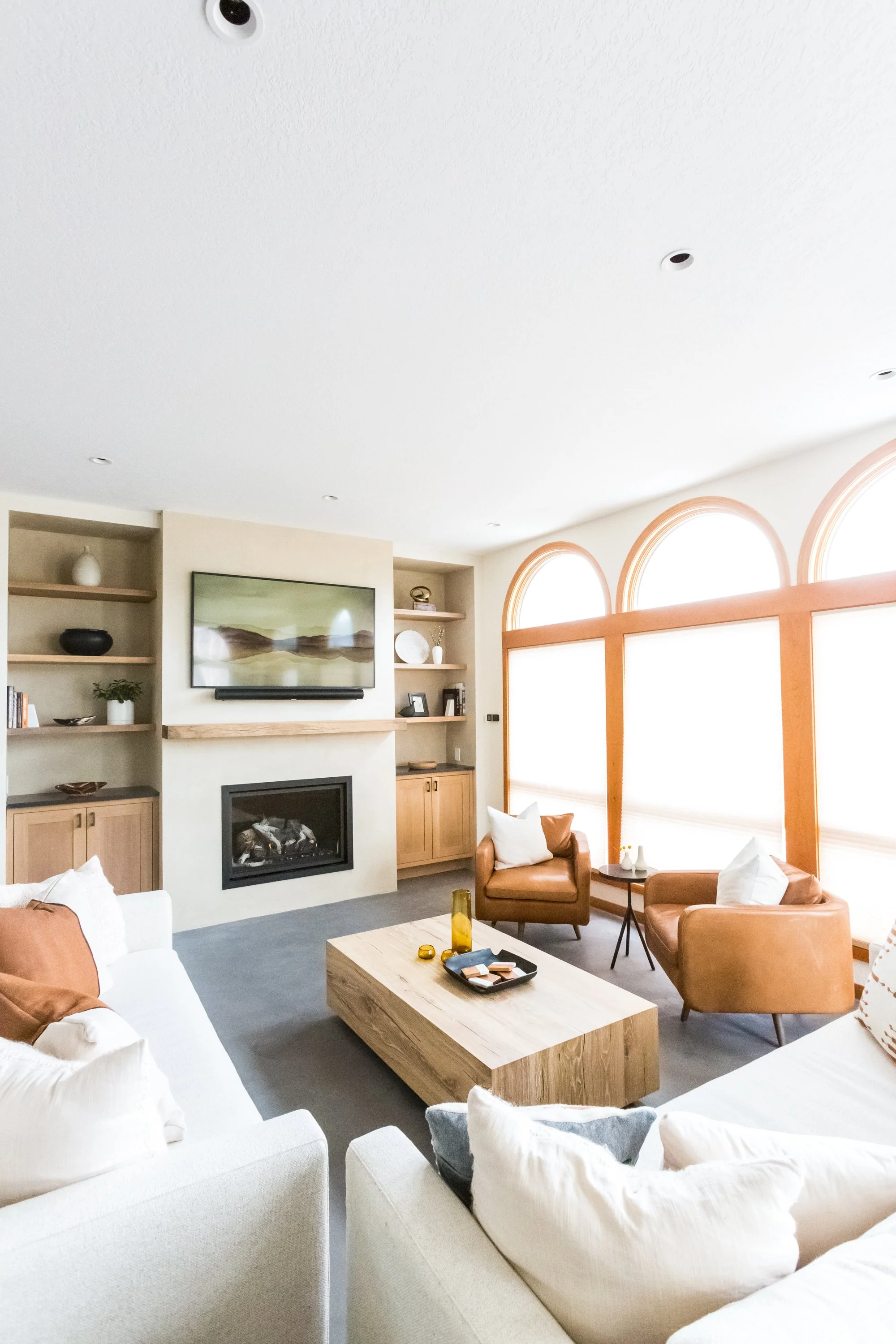 Living room with a fireplace, wall-mounted TV, beige built-in shelves, arched windows, and tan leather chairs.