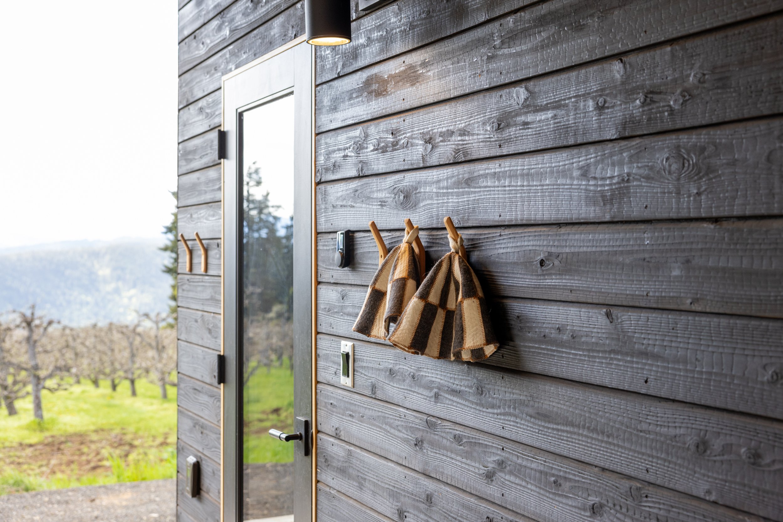 A wooden exterior wall with a mirror, a black outdoor light, a doorbell, a switch, and clothes hooks holding two checkered cloths, with a scenic landscape seen through the window.