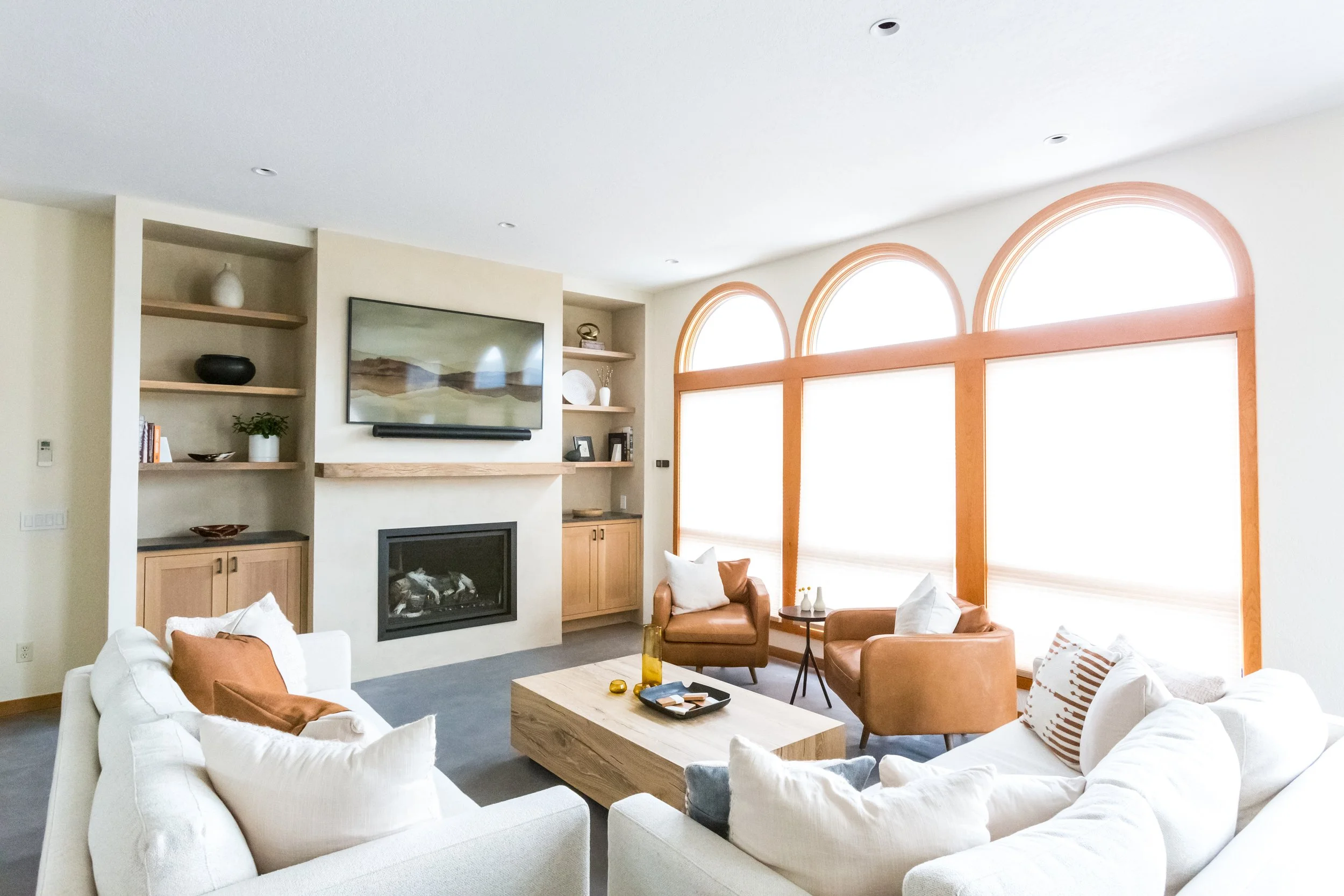 A spacious living room with large arched windows, a white sofa, tan armchairs, a wooden coffee table, a TV mounted above a fireplace, and built-in shelves with decorative items.