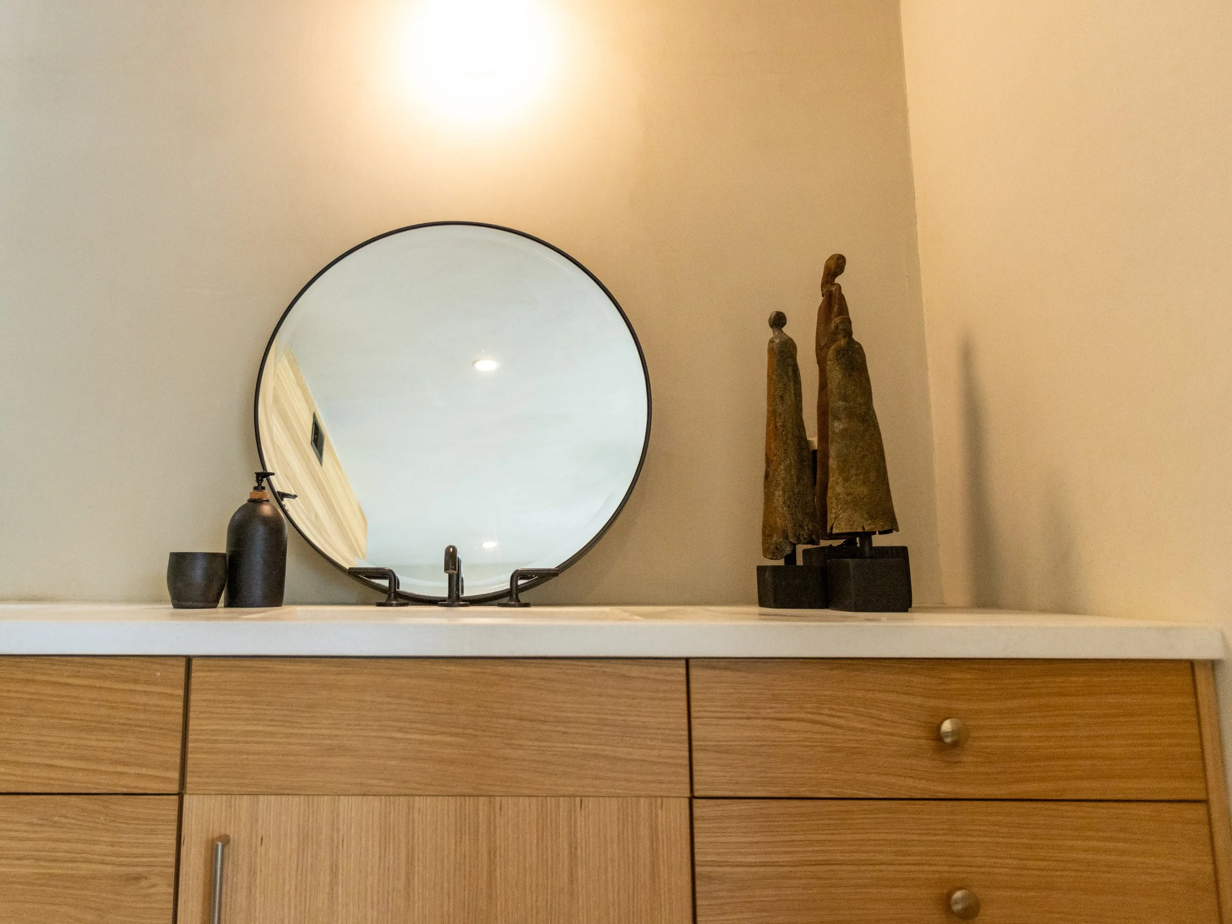 A bathroom vanity with a round mirror, two black vases, and three wooden sculptures on a black stand, above wooden drawers.