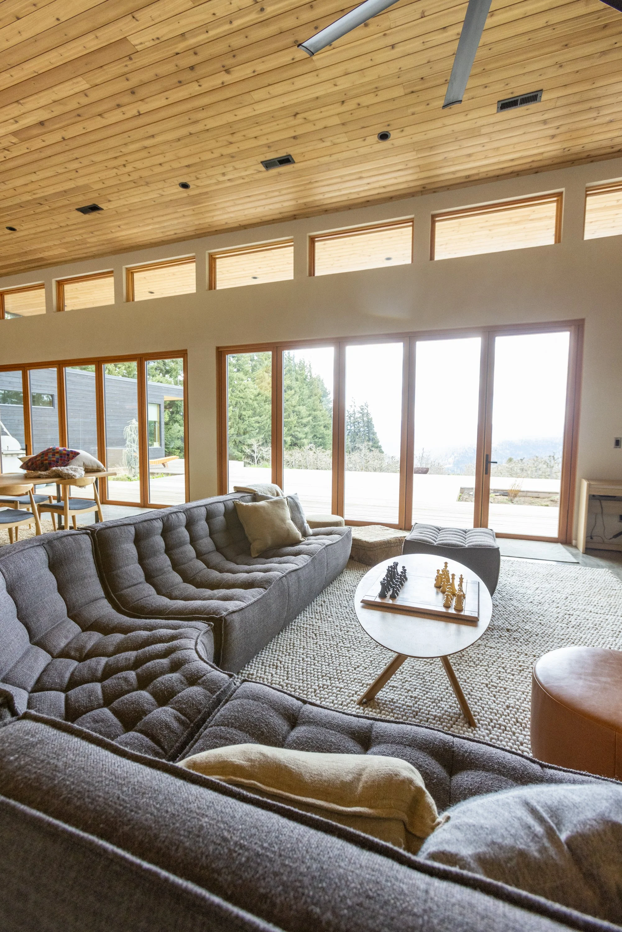 Living room with large gray sectional sofa, round coffee table with chess set, beige ottoman, and a rug. Floor-to-ceiling glass doors and windows reveal a deck and greenery outside. Wooden ceiling with beams and small high windows.