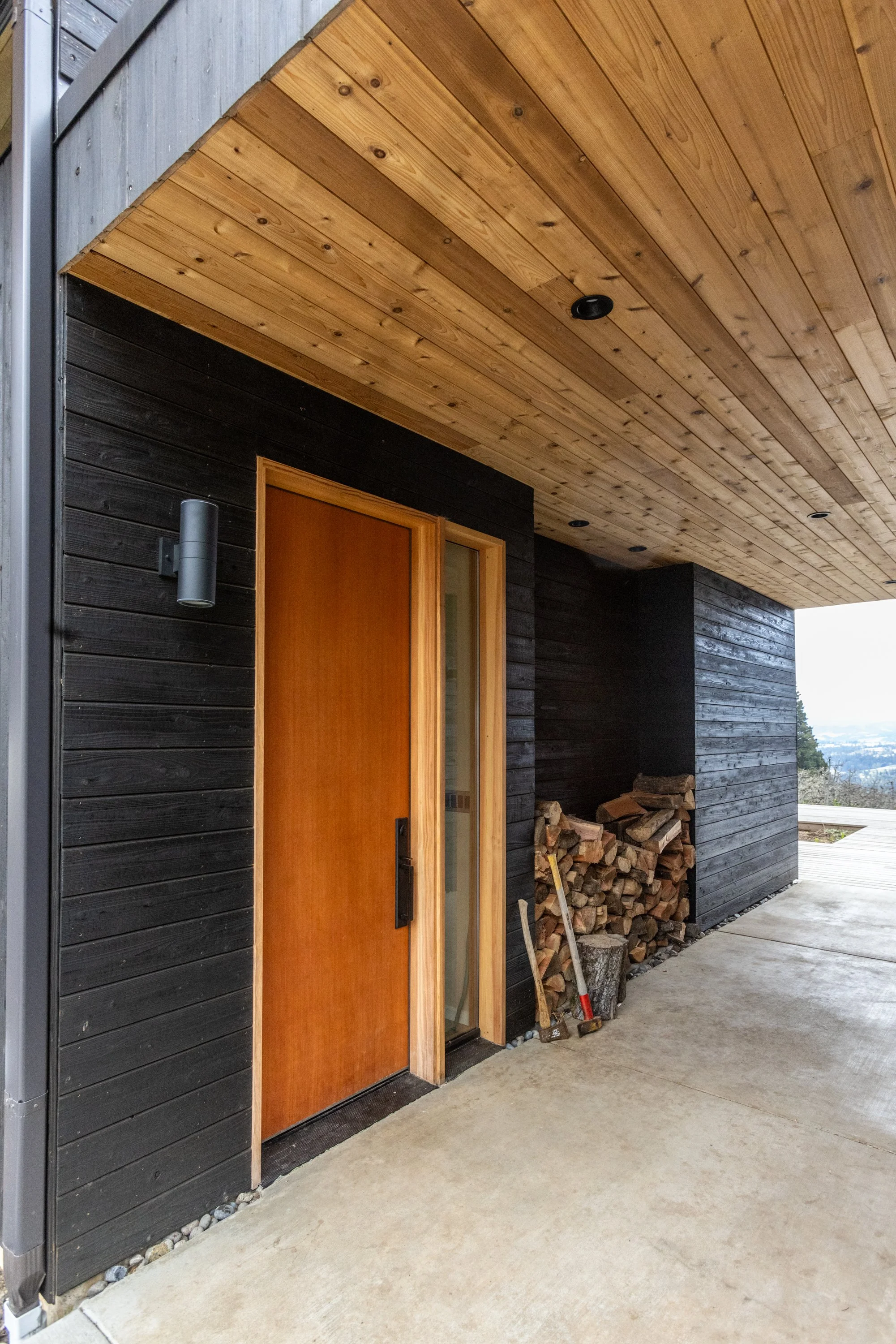 Modern house exterior with black wooden siding, a wooden front door, a woodpile next to the house, and a concrete walkway.