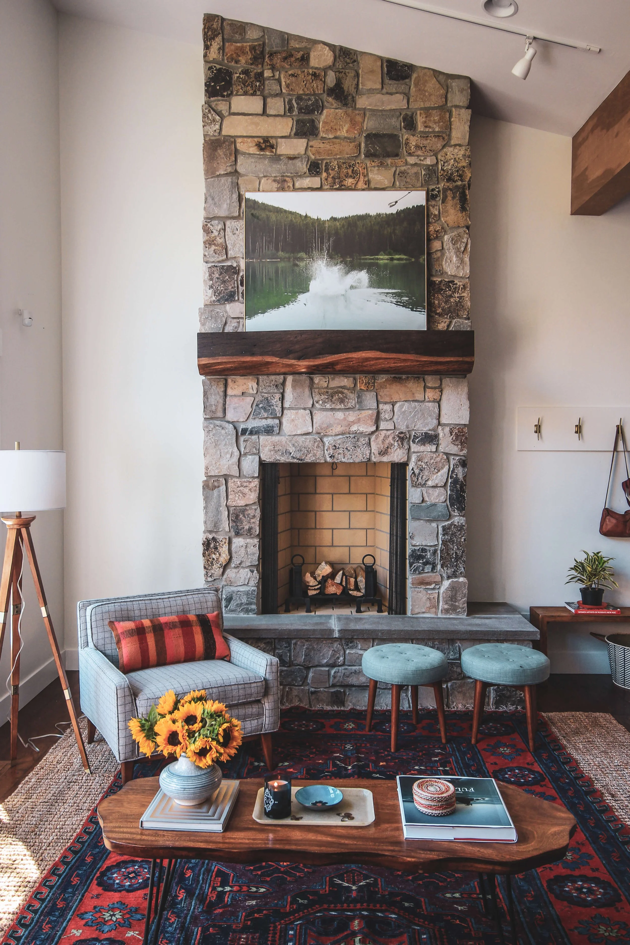Living room with stone fireplace, two teal stools, a wooden coffee table with decorative items including a sunflower bouquet, a book, a candle, and a decorative box, a plaid-patterned armchair with a pillow, a rug, a side table with a book and a box,