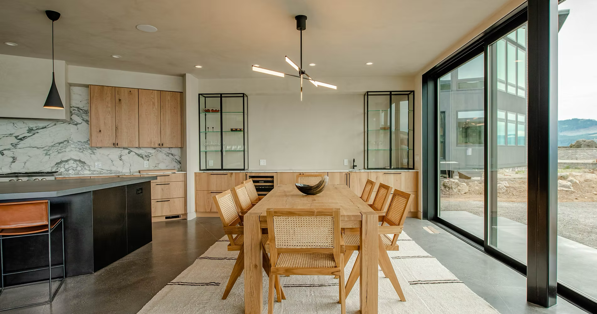 Modern kitchen and dining area with large glass doors overlooking a rocky landscape, featuring wooden dining table with eight matching chairs, glass display cabinets, marble backsplash, black and wooden kitchen cabinetry, and minimalistic lighting fi