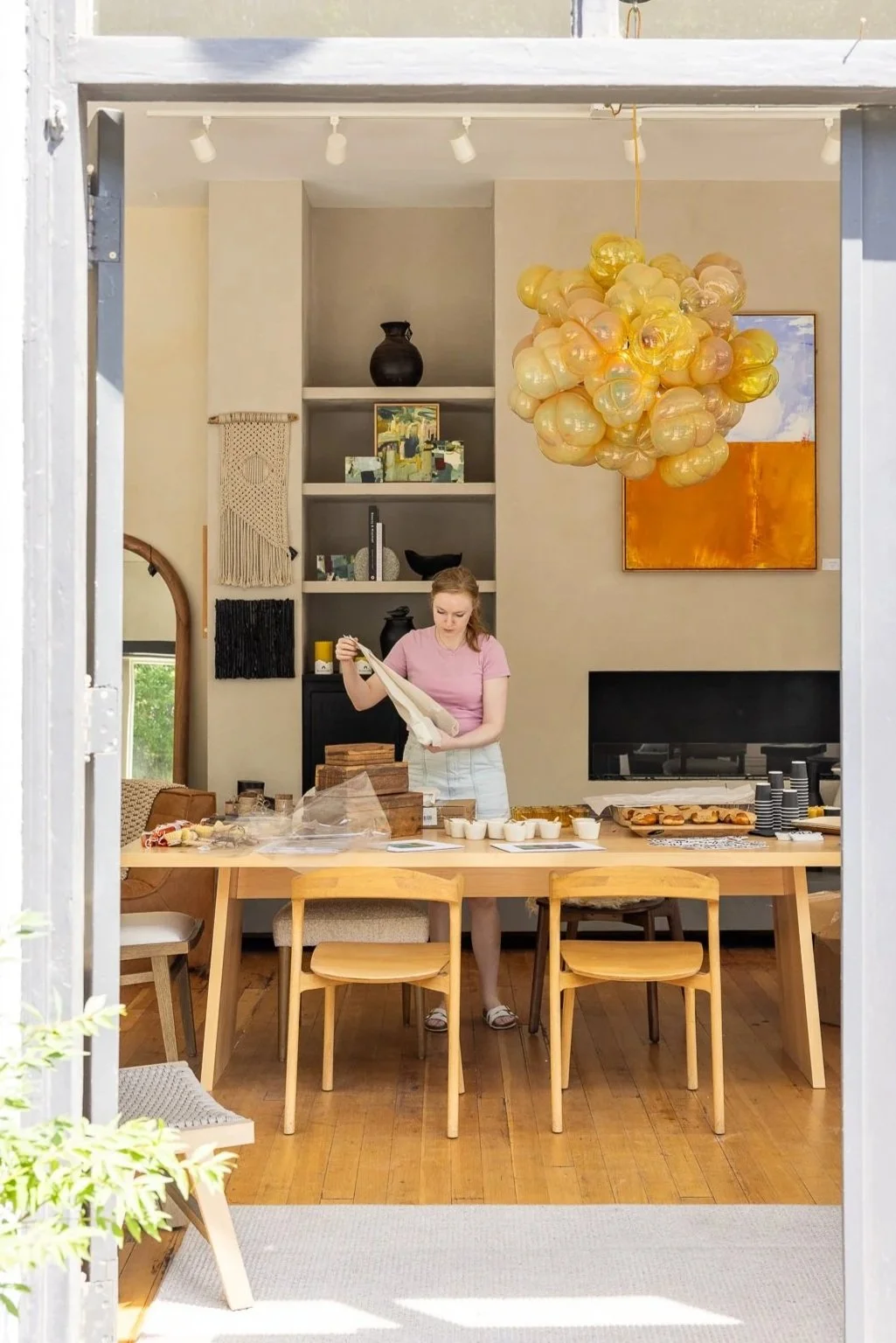 A woman in a pink shirt and white shorts preparing food at a dining table set with plates and snacks in a well-lit, modern interior with artwork, a built-in fireplace, and decorative shelving.