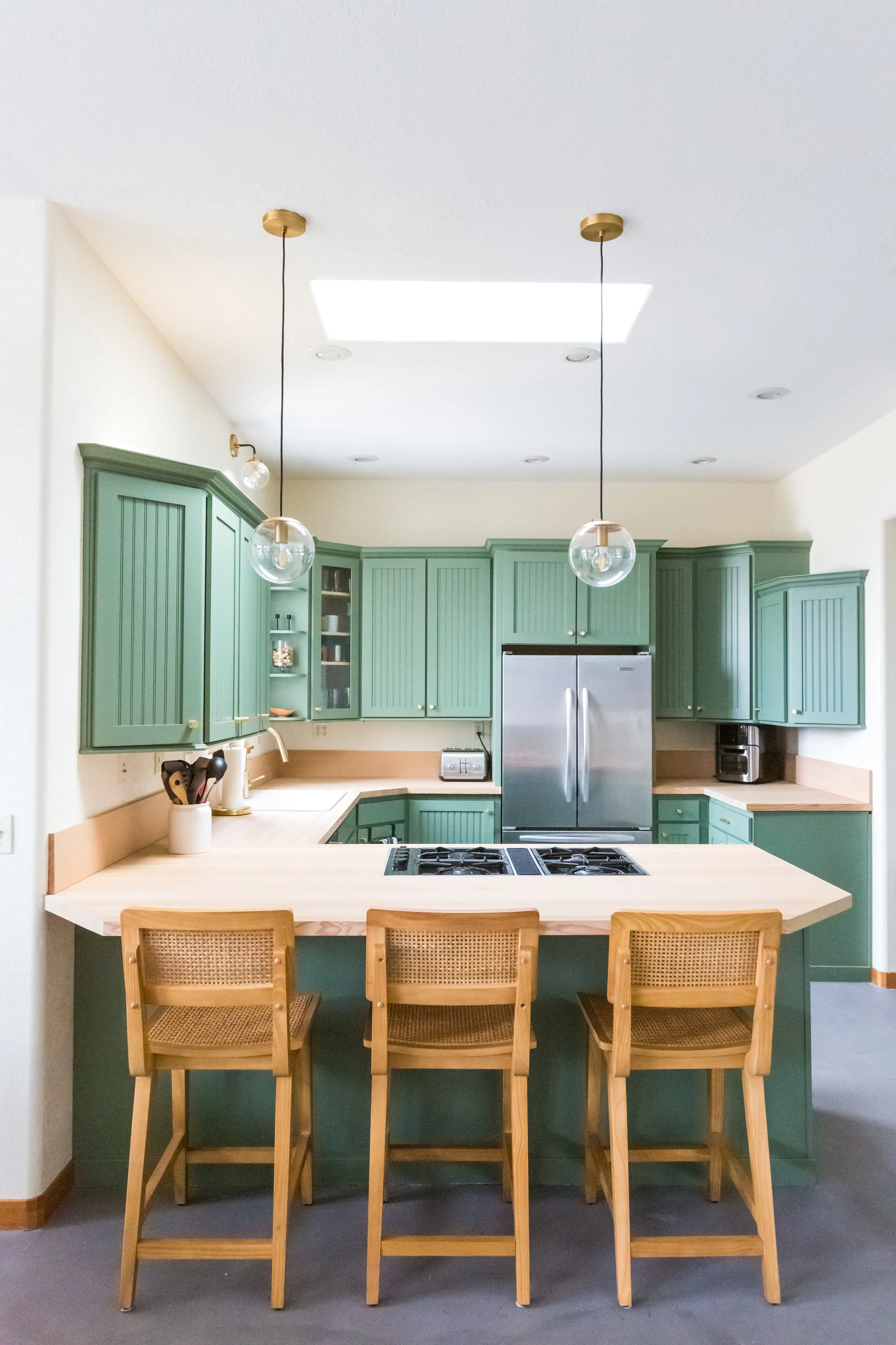 A kitchen with green cabinets, a beige countertop, a stainless steel refrigerator, and hanging pendant lights with globe bulbs above a breakfast bar with four wooden chairs. There is a skylight on the ceiling.