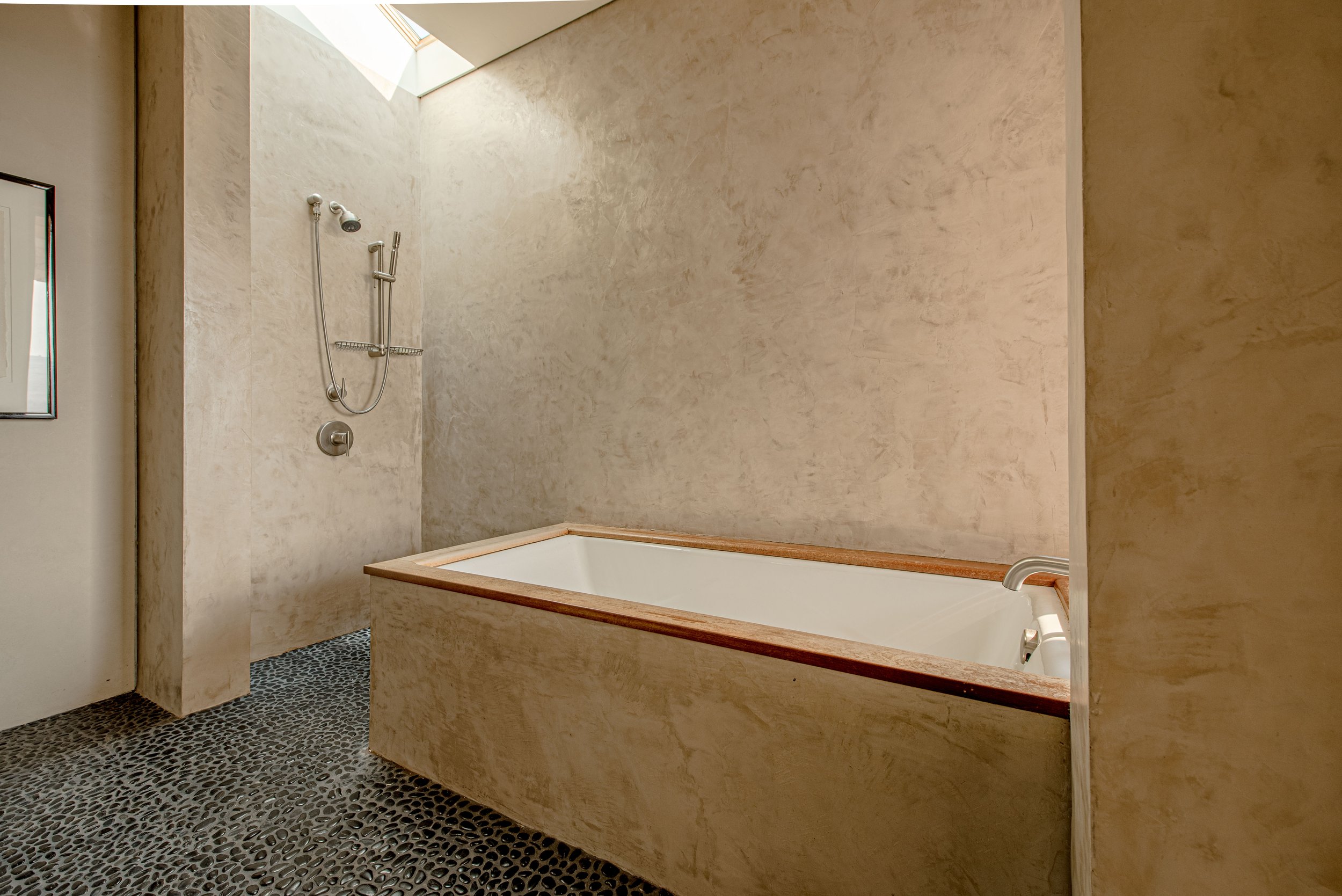 A bathroom with a beige textured wall, a bathtub with a wooden ledge, a shower with a handheld spray, and a skylight providing natural light.