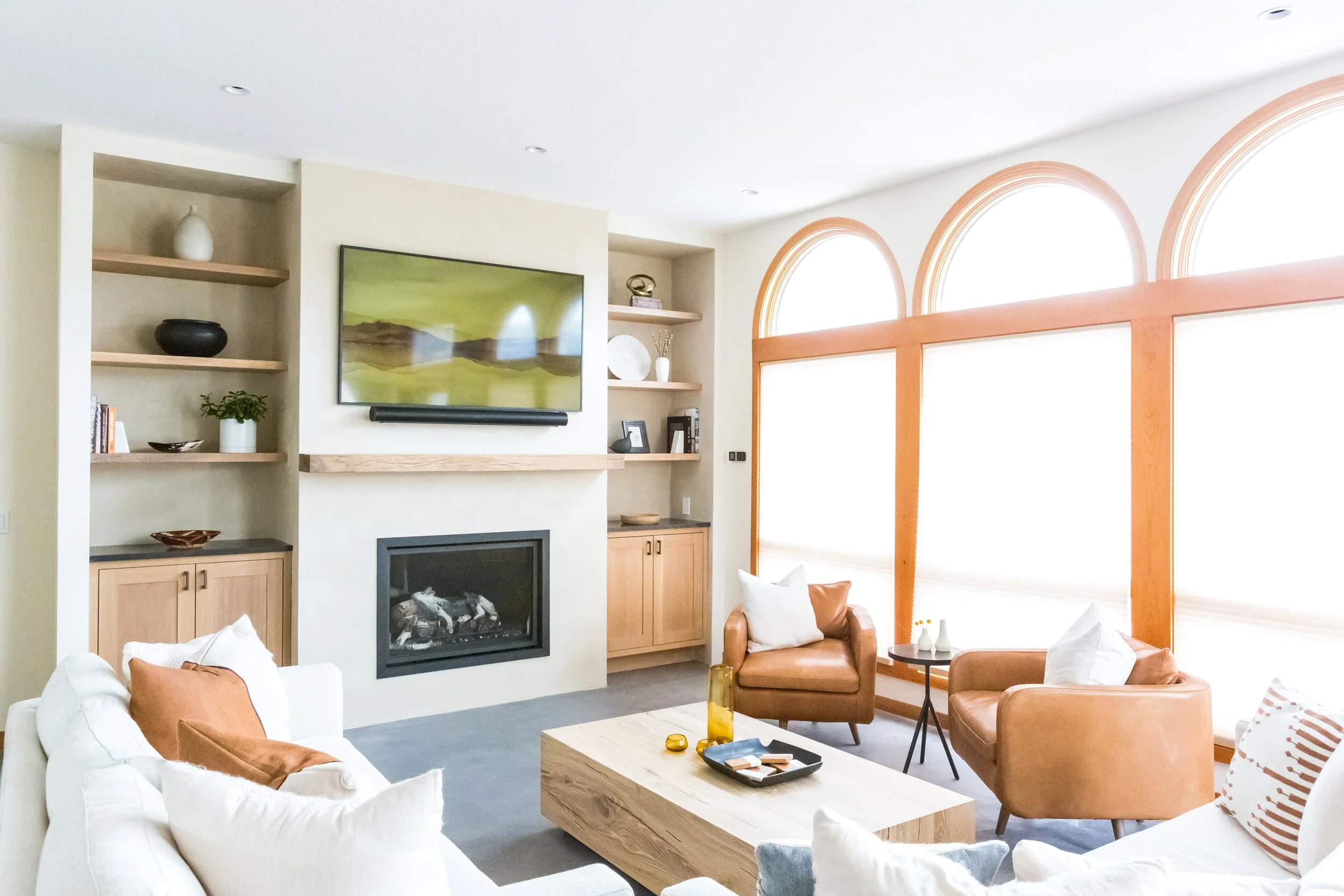 Living room with white sectional sofa, tan leather chairs, wooden coffee table, built-in shelves with decor, wall-mounted TV showing a landscape, fireplace, large arched windows with wooden trim, and various cushions and decor items.