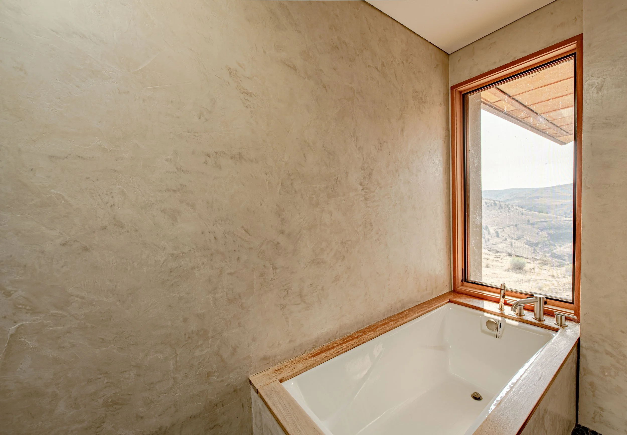 Bathroom with a large window overlooking a scenic landscape, featuring a bathtub with a wooden frame, a silver faucet, and beige textured walls.