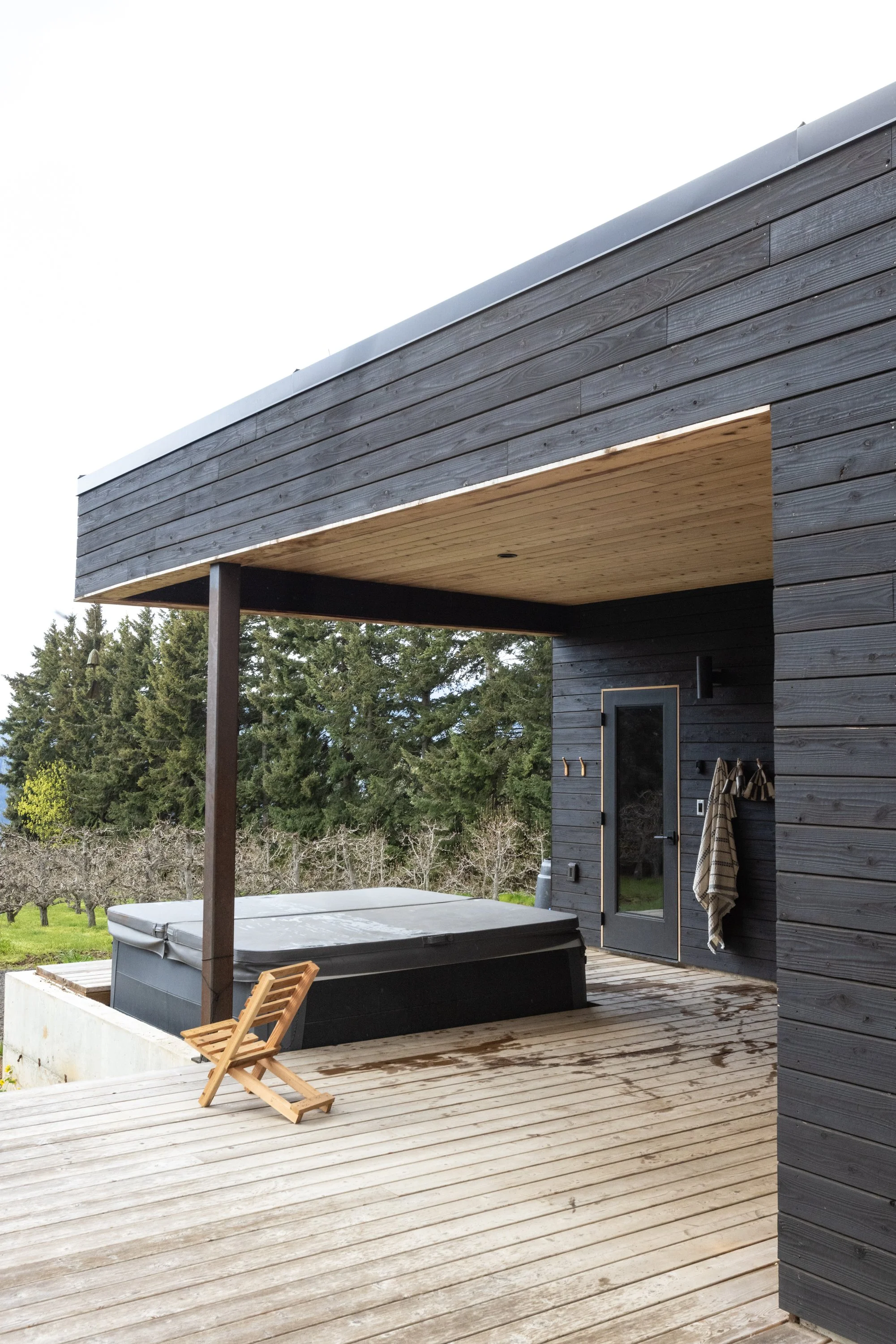 A modern house porch with black wooden walls, a wooden ceiling, and a hot tub on a wooden deck. A small wooden folding chair is placed nearby, and there are trees in the background.