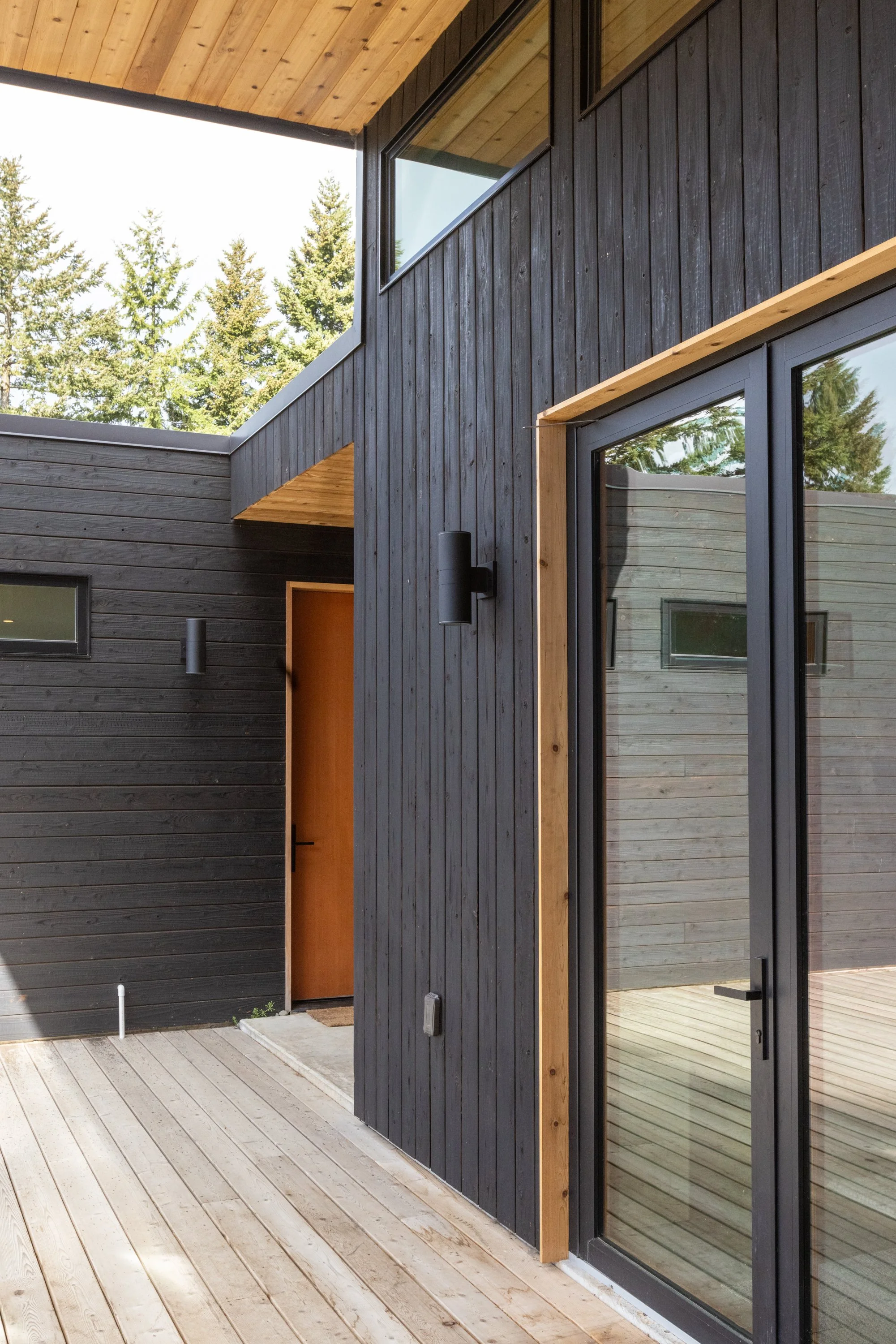 Modern house exterior with black wooden siding, large glass sliding door, wooden trim, and a wooden ceiling over a wooden deck, with trees visible through the windows.