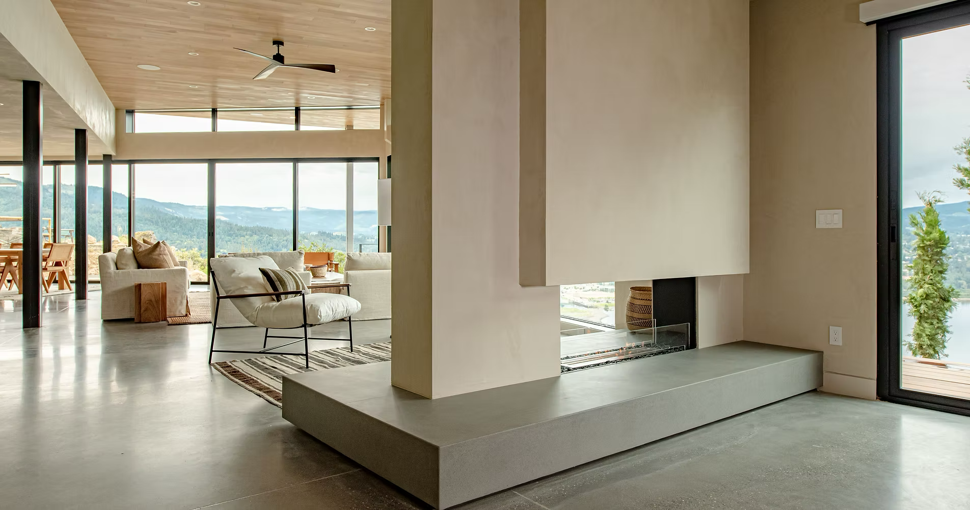 Modern living room with large glass windows showing mountain views, a white sofa, a black metal chair with white cushion, a fireplace, and a ceiling fan.