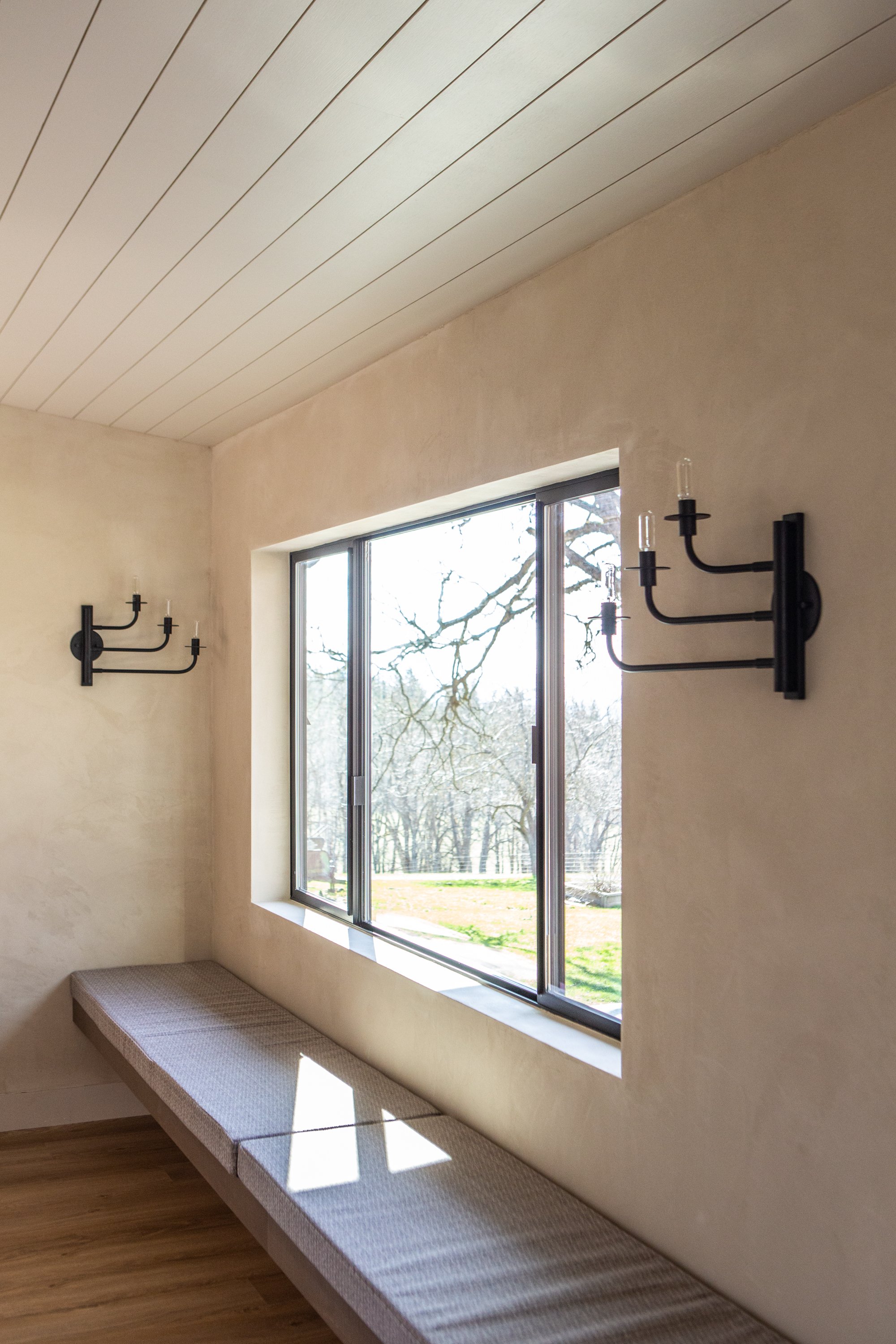 A window with a scenic outdoor view of trees and grass. Inside, there are black sconces with candle-shaped light bulbs on the beige wall. A cushioned bench runs along the wall beneath the window, and the room has wood flooring.