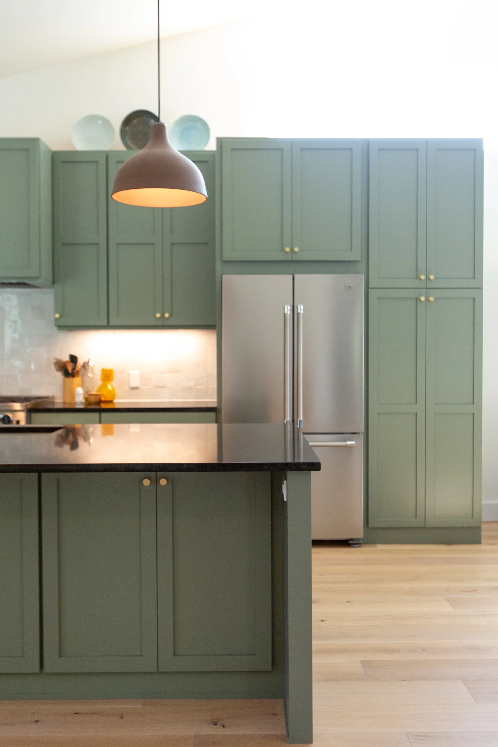 A kitchen with green cabinets, a stainless steel refrigerator, and a wooden floor. There are three ceramic plates on top of the cabinets and a black pendant light hanging from the ceiling. On the counter, there are yellow condiments and utensils.