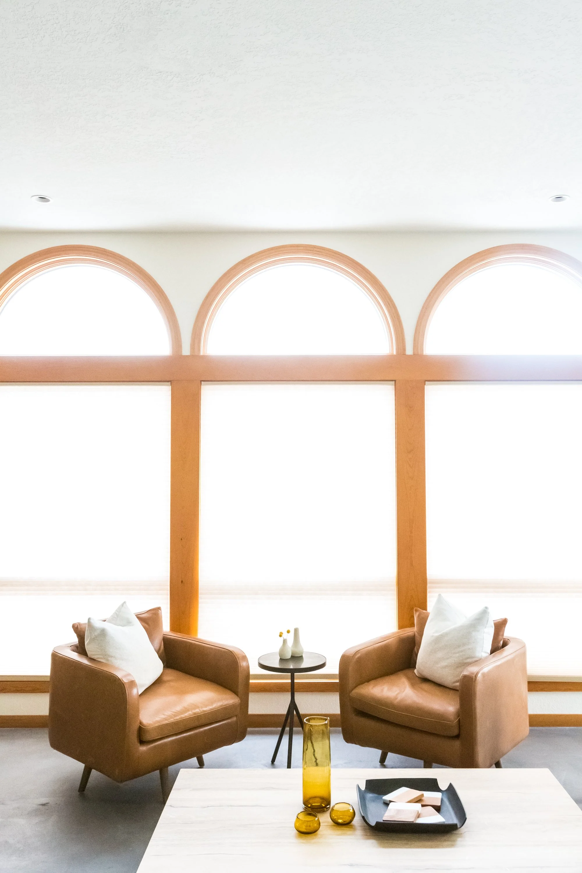 A cozy seating area with two tan leather armchairs with white pillows, a small round black side table with decorative vases, and large arched windows with wooden frames allowing bright natural light into the room.