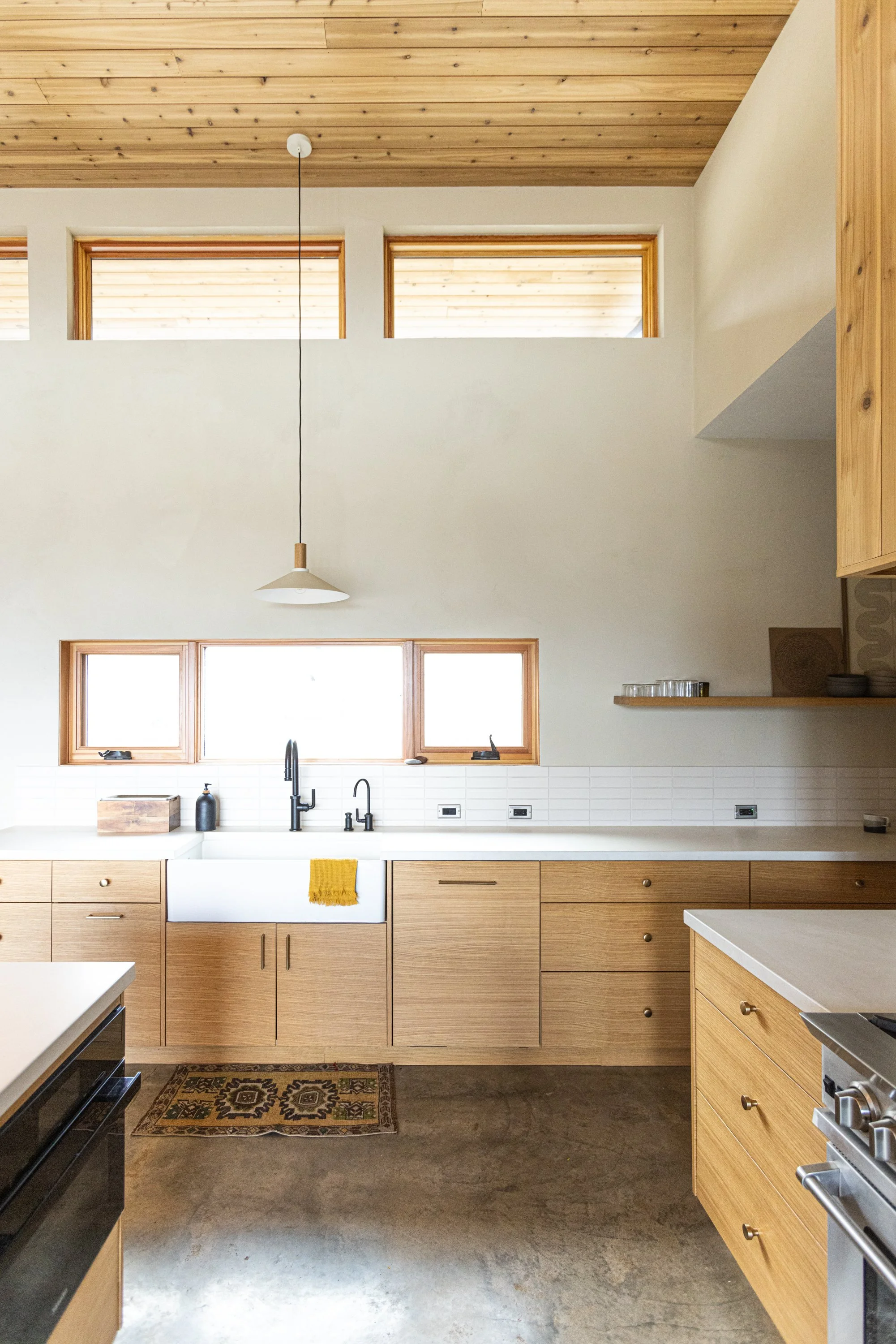 Modern kitchen with light wood cabinets, white countertop, black sink, and a window with natural light. Wooden ceiling with recessed lighting, hanging light fixture, and a patterned rug on the floor.