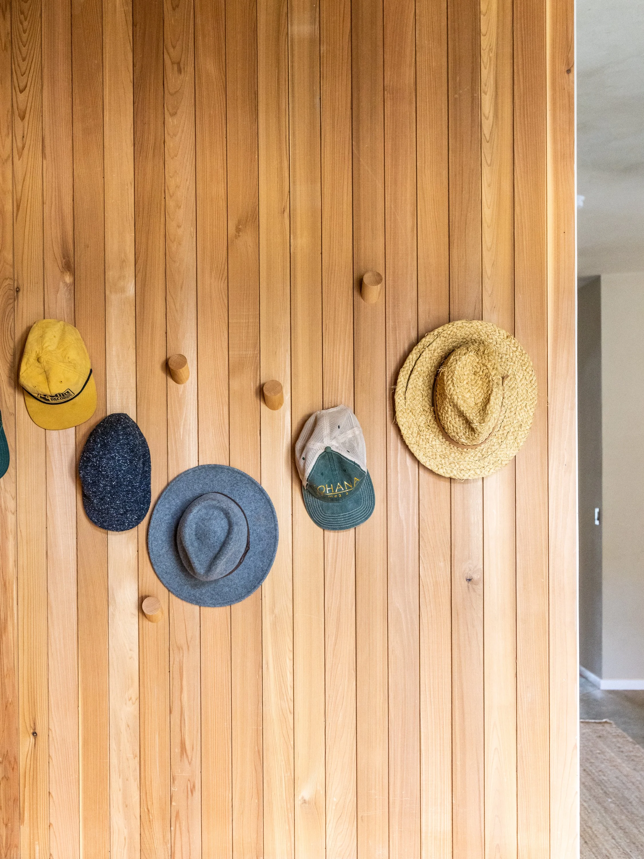 Wood-paneled wall with six hooks holding hats and caps, including a yellow cap, grayish-blue wide-brim hat, green and beige cap, and two straw hats.