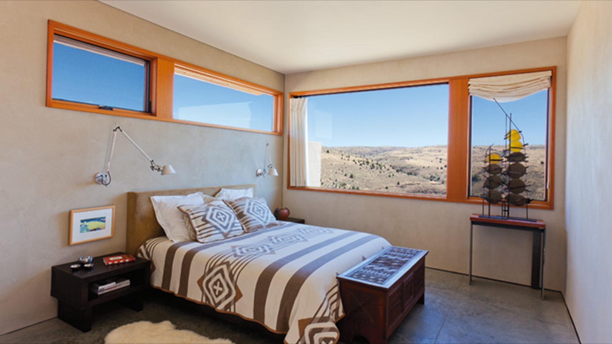 Bedroom with large windows showing a desert landscape, bed with geometric patterned bedding, wall-mounted lamps, small framed picture, decorative ship model, and side table with books and remote controls.