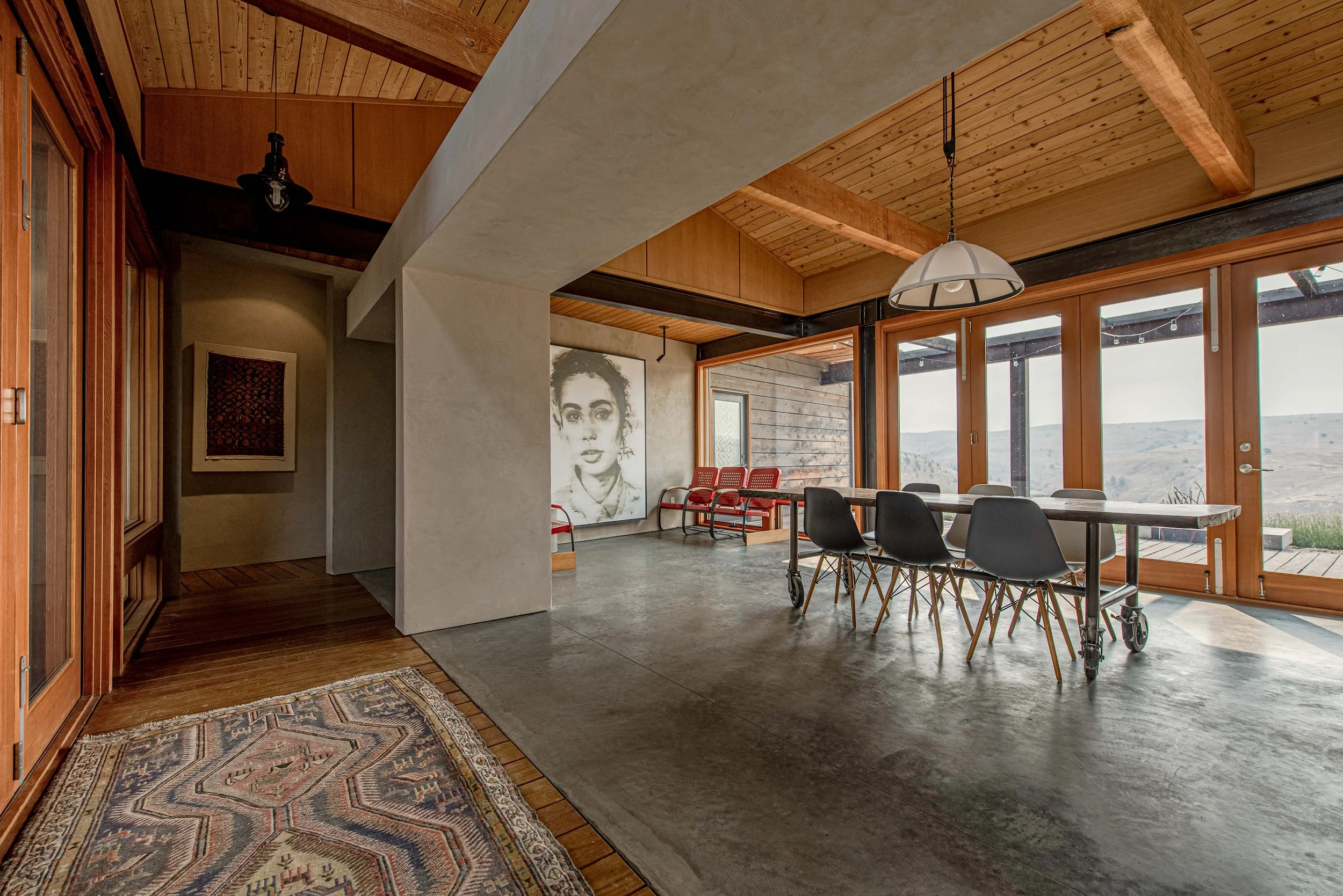 Interior of a modern house with wooden ceiling beams, large glass windows, and a dining area with chairs around a table. There are artworks on the walls, including a black-and-white portrait of a woman and a colorful framed piece. A patterned rug is 