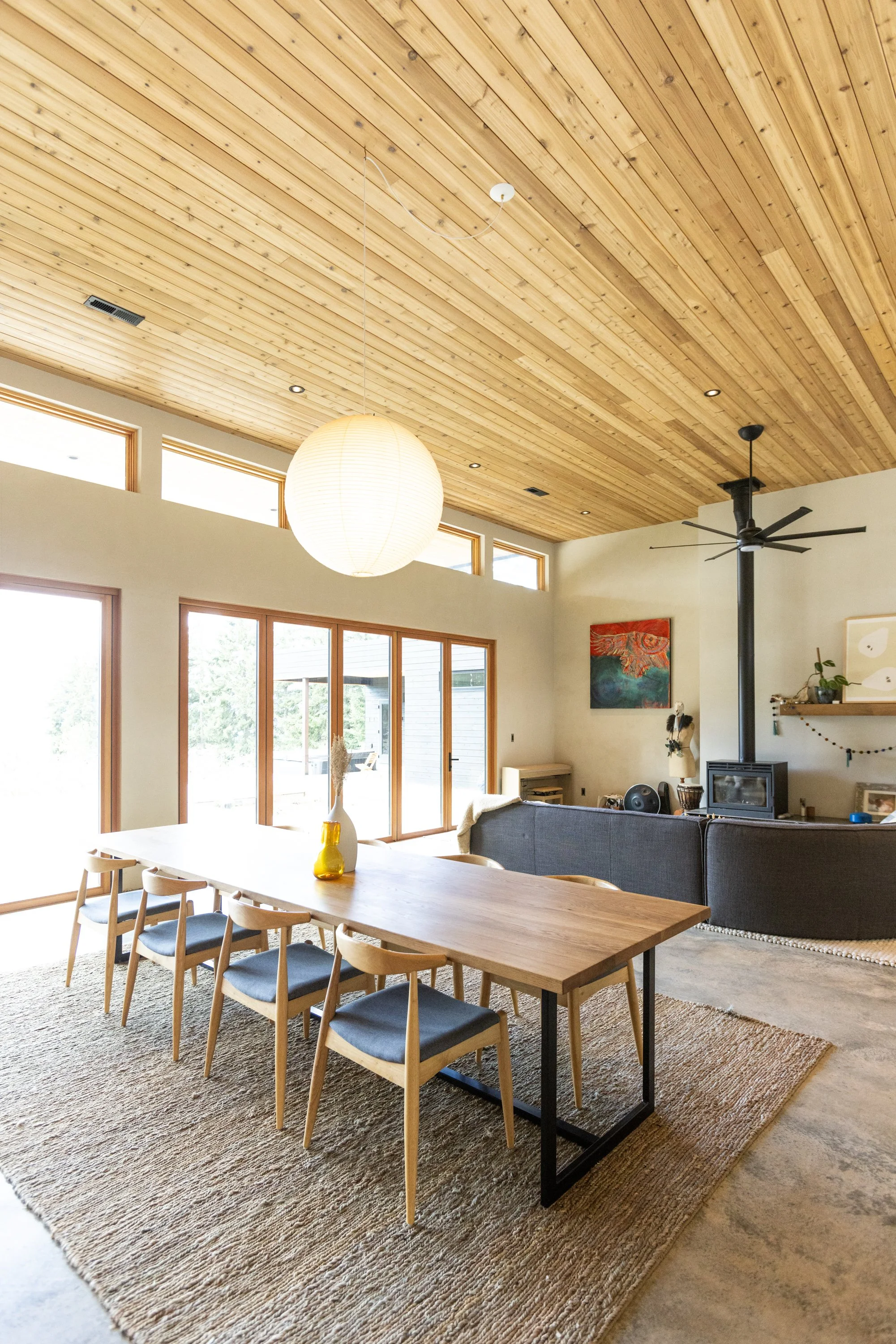Open-concept living and dining area with natural wood ceiling, large windows, a dining table with six chairs, a white paper lantern pendant light, a gray sofa, a wood-burning stove, green plant, wall art, and a small shelf with decorative items.
