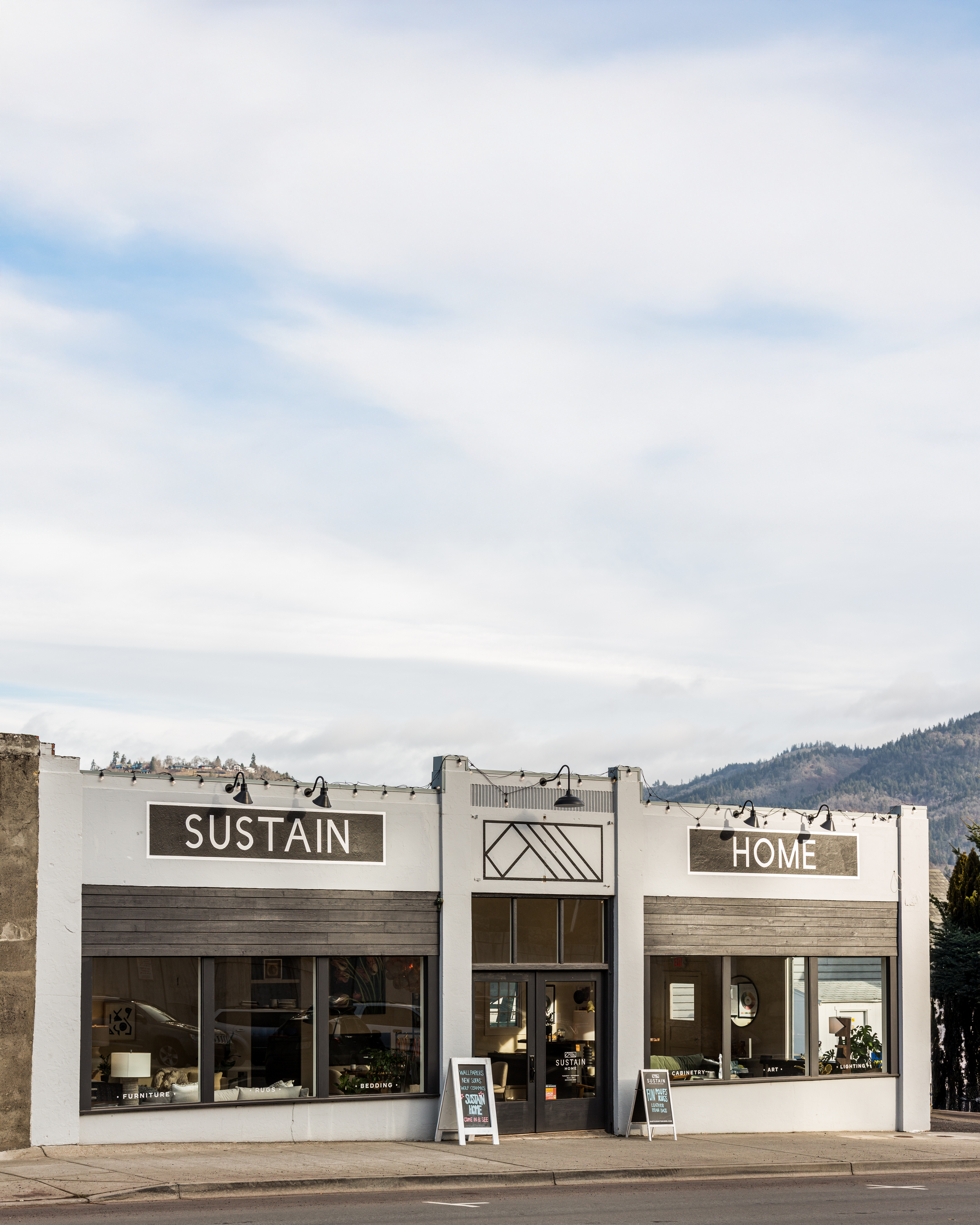 A storefront with large windows displaying home décor items such as furniture, bedding, rugs, and art. Signs above read 'SUSTAIN' and 'HOME'. Small chalkboard signs outside advertise a workshop event and available home furnishings.