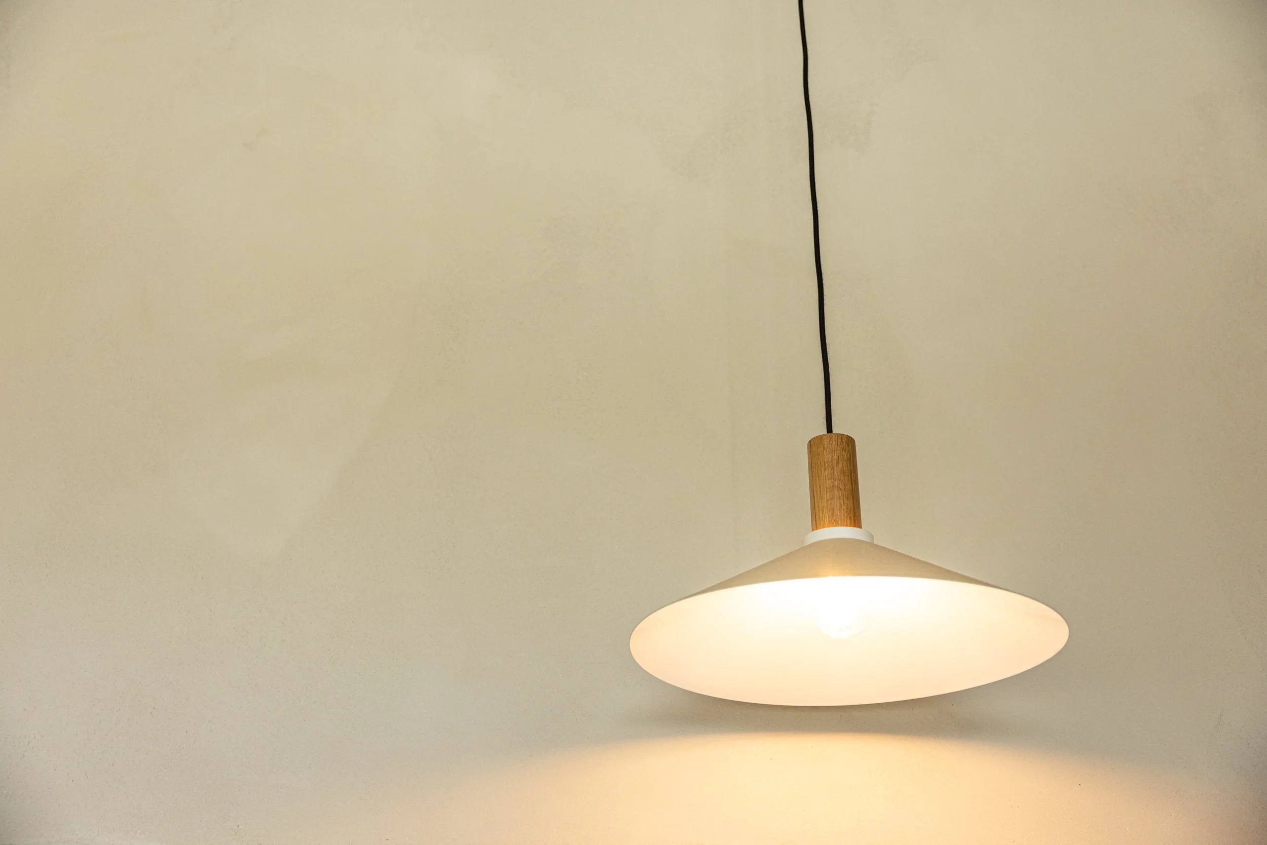 A ceiling light fixture with a white shade, a black cord, and a wooden element attached to the shade, against a plain beige ceiling.