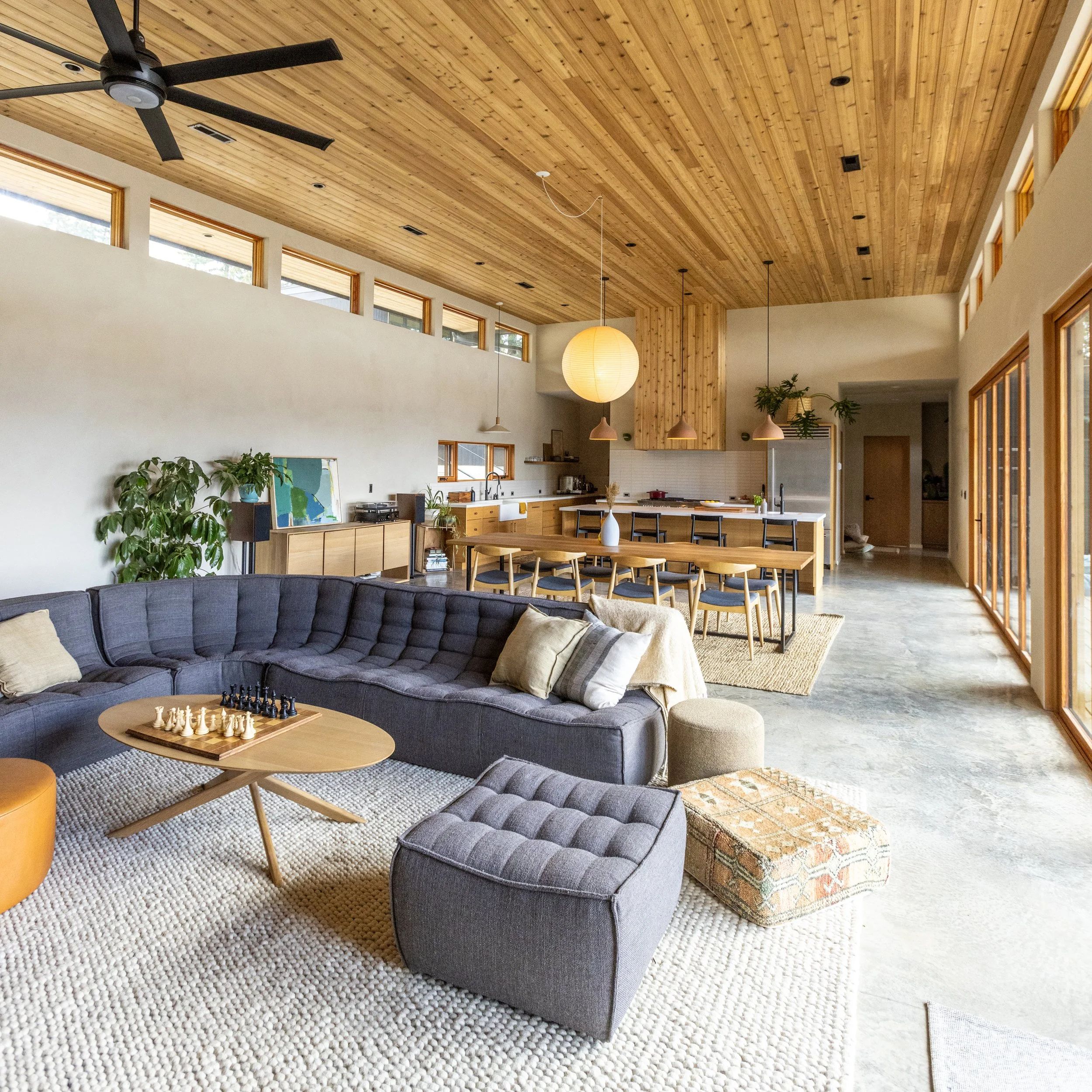 A living room with a dark gray curved sofa on a textured white rug, a wooden coffee table with a chessboard, and several decorative pillows. The room has a high wooden ceiling with a ceiling fan, large windows, and pendant lights over a dining area w
