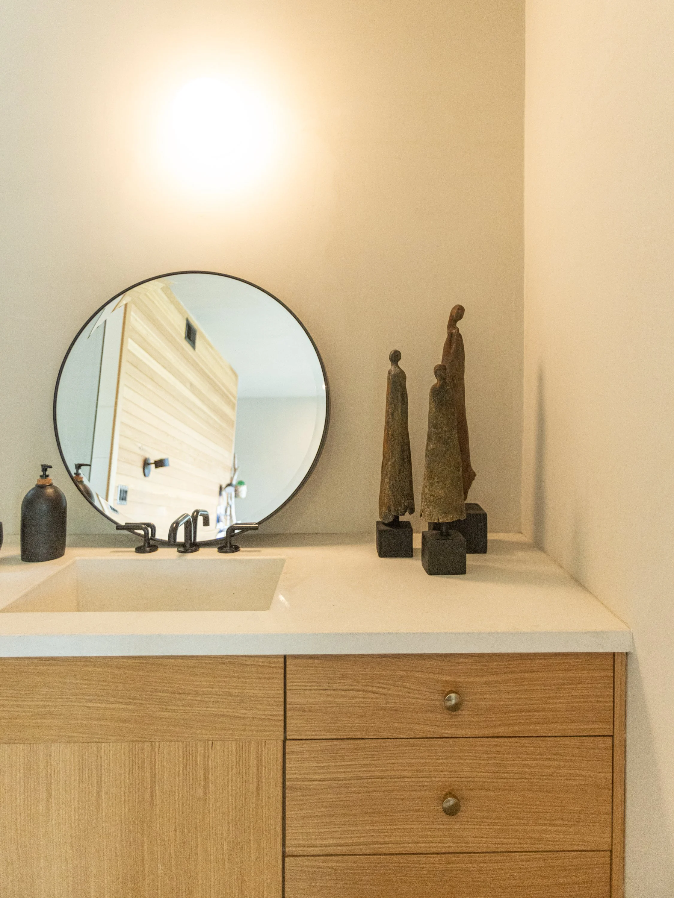 A bathroom vanity with a round mirror, a sink, a soap dispenser, and three tall decorative statues on the counter.