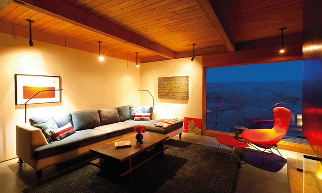Cozy living room with a gray sofa, red and patterned pillows, a wooden coffee table with a flower vase and books, artwork on the walls, a large window showing a night sky, and a red lounge chair with an ottoman.
