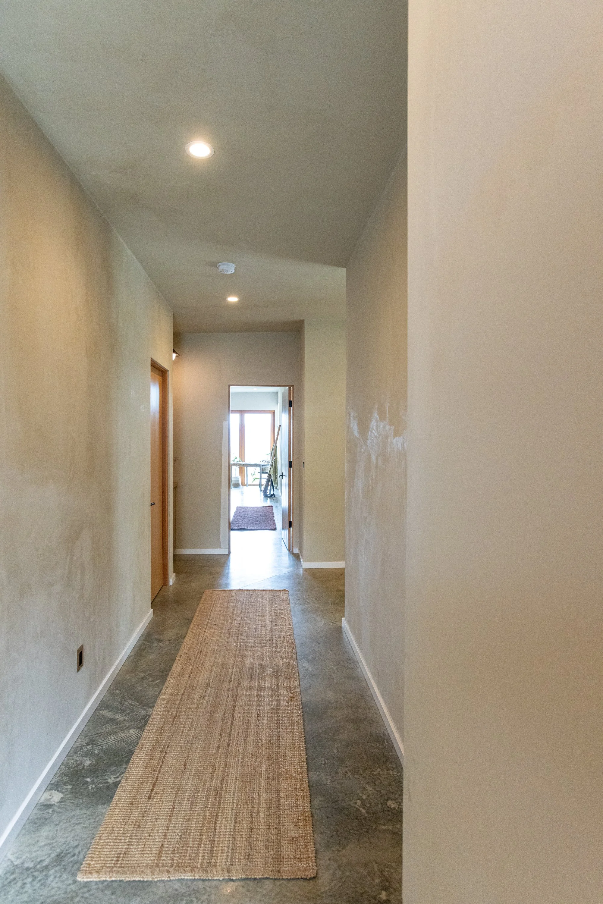 A hallway with beige walls, a beige rug on a gray marble floor, and a doorway at the end leading to a room with large windows and a balcony.