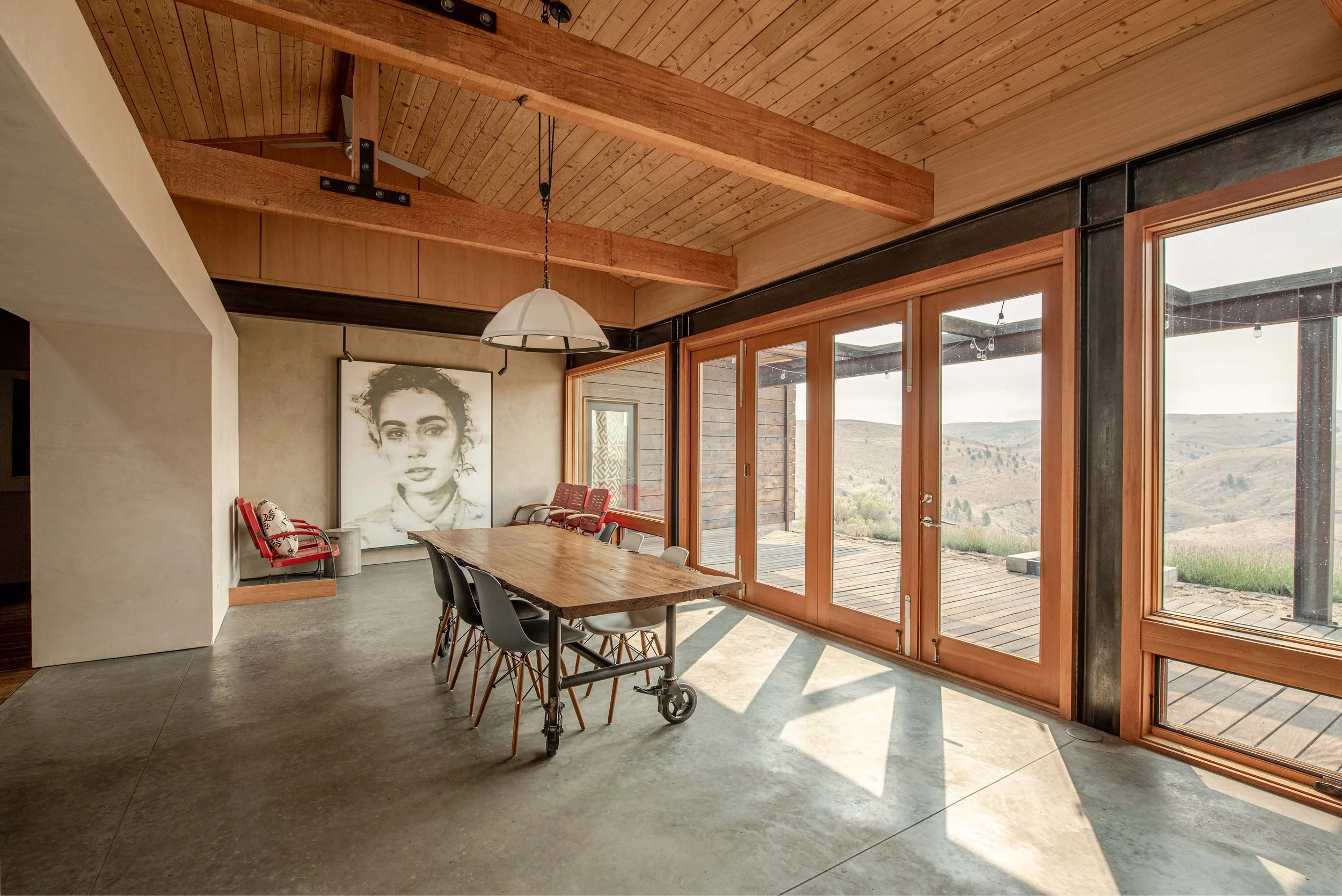 Modern dining room with large floor-to-ceiling windows overlooking a mountain landscape, wooden ceiling with exposed beams, a large wooden table, black chairs, a pendant light, and artwork on the wall.