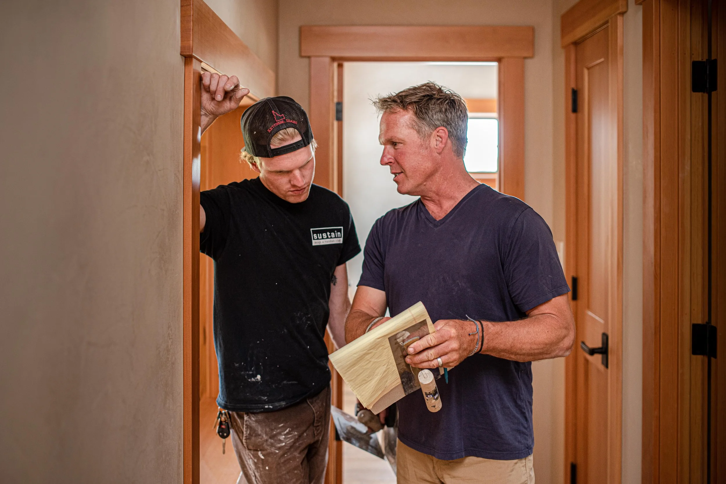 Two men engaged in a discussion inside a hallway with wooden doors, one holding a piece of wood and a small container, the other listening closely.