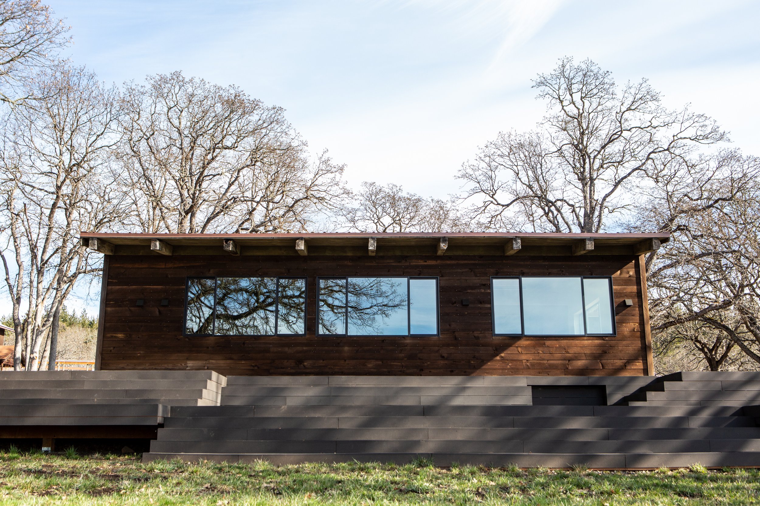 A modern house with dark wooden exterior and large glass windows, surrounded by leafless trees and a grassy lawn under a partly cloudy sky.