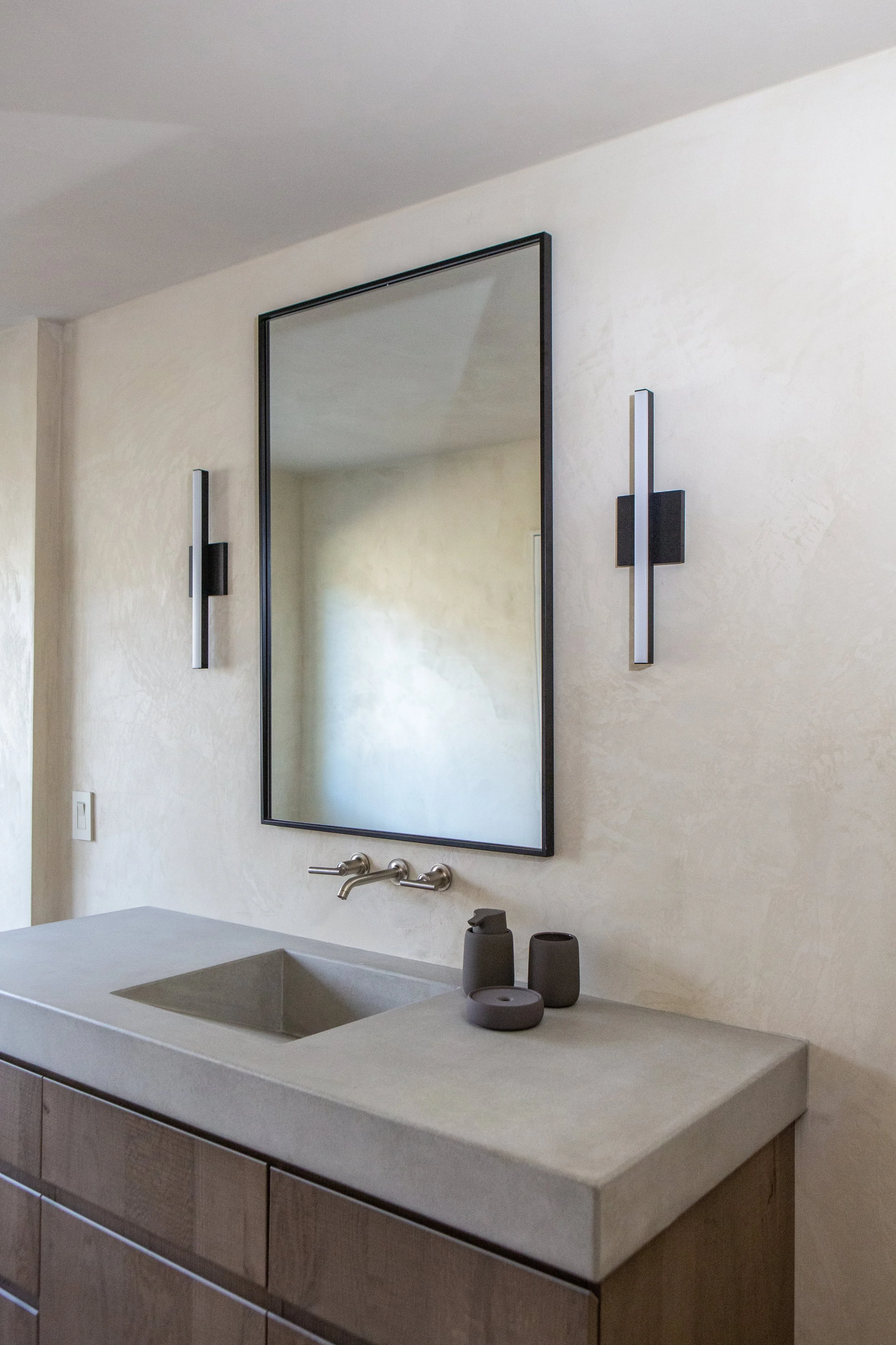 Modern bathroom with a rectangular mirror, two vertical black and white sconces, a concrete vanity with a built-in sink, and decorative dark gray vases on the countertop.