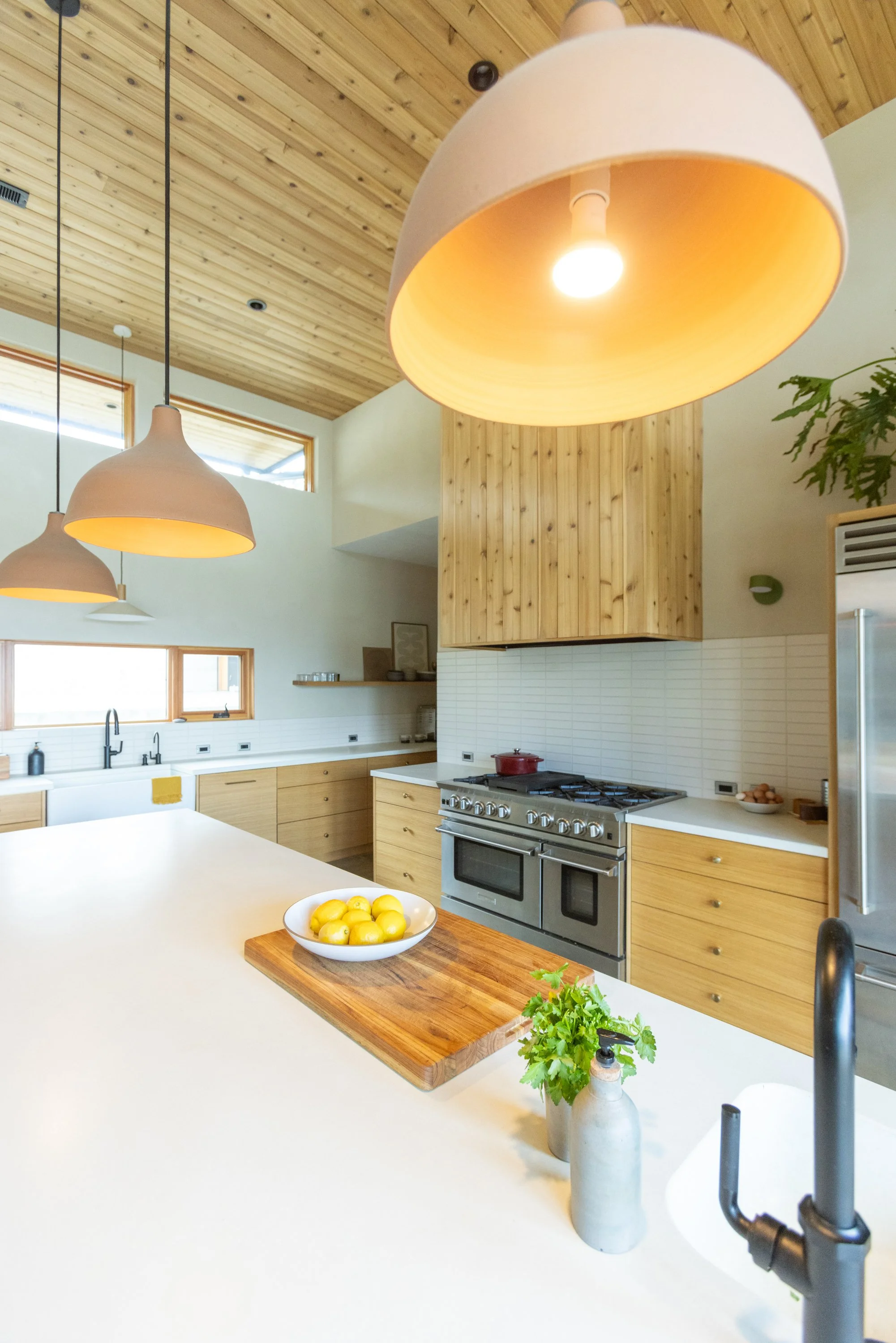 Modern kitchen with wooden cabinets and ceiling, large white island countertop, bowl of lemons on a wooden cutting board, small green plant, and pendant lights.