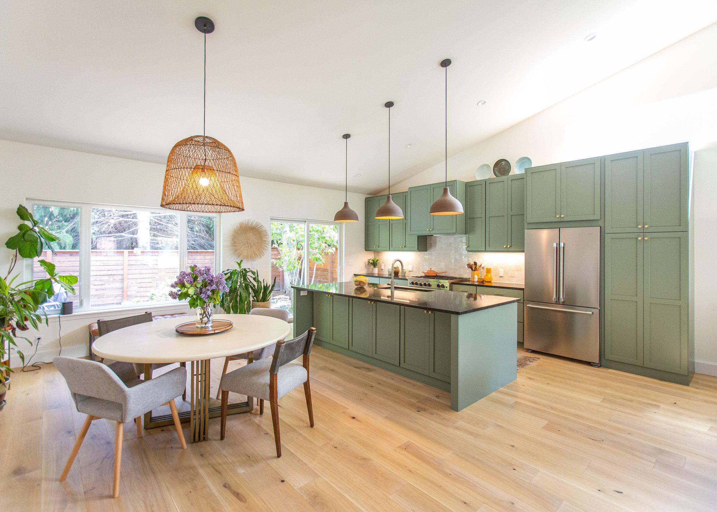 A kitchen with green cabinets, a black countertop island, stainless steel refrigerator, three pendant lights, a round dining table with four chairs, plants, and large windows overlooking a backyard with trees and a wooden fence.