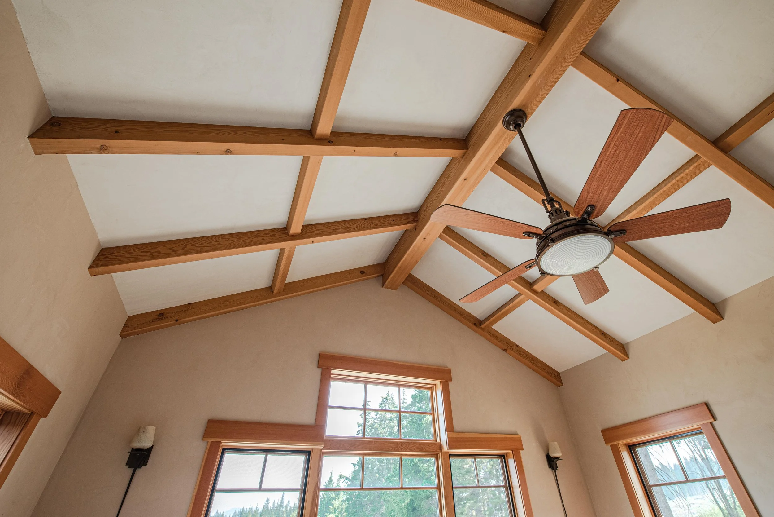 High vaulted ceiling with exposed wooden beams and a ceiling fan with wooden blades and a glass light cover. Large windows with wooden trim allow natural light into the room.
